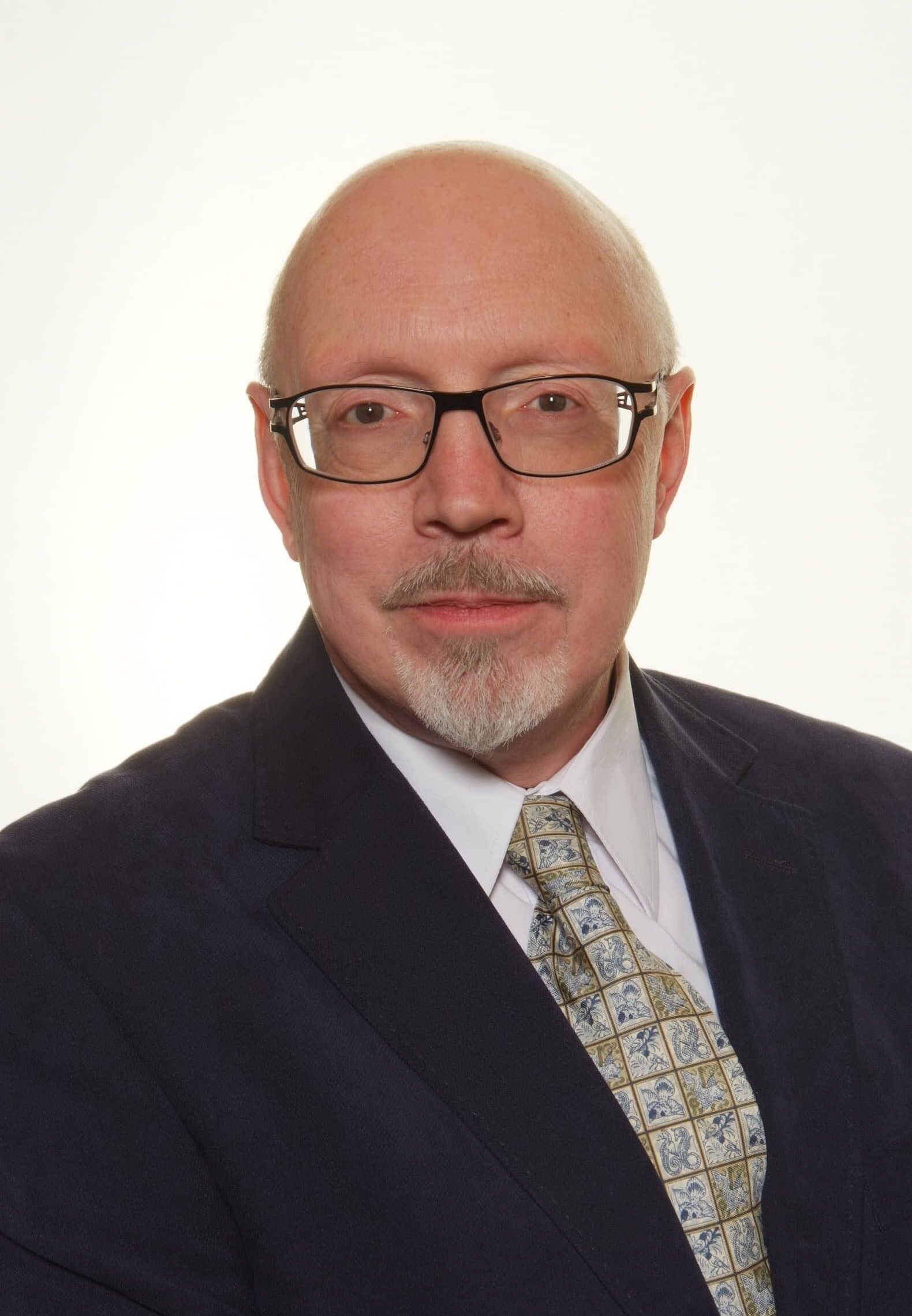 A middle-aged man with glasses, a goatee, and a receding hairline, wearing a dark suit, light shirt, and patterned tie, poses against a plain light background.