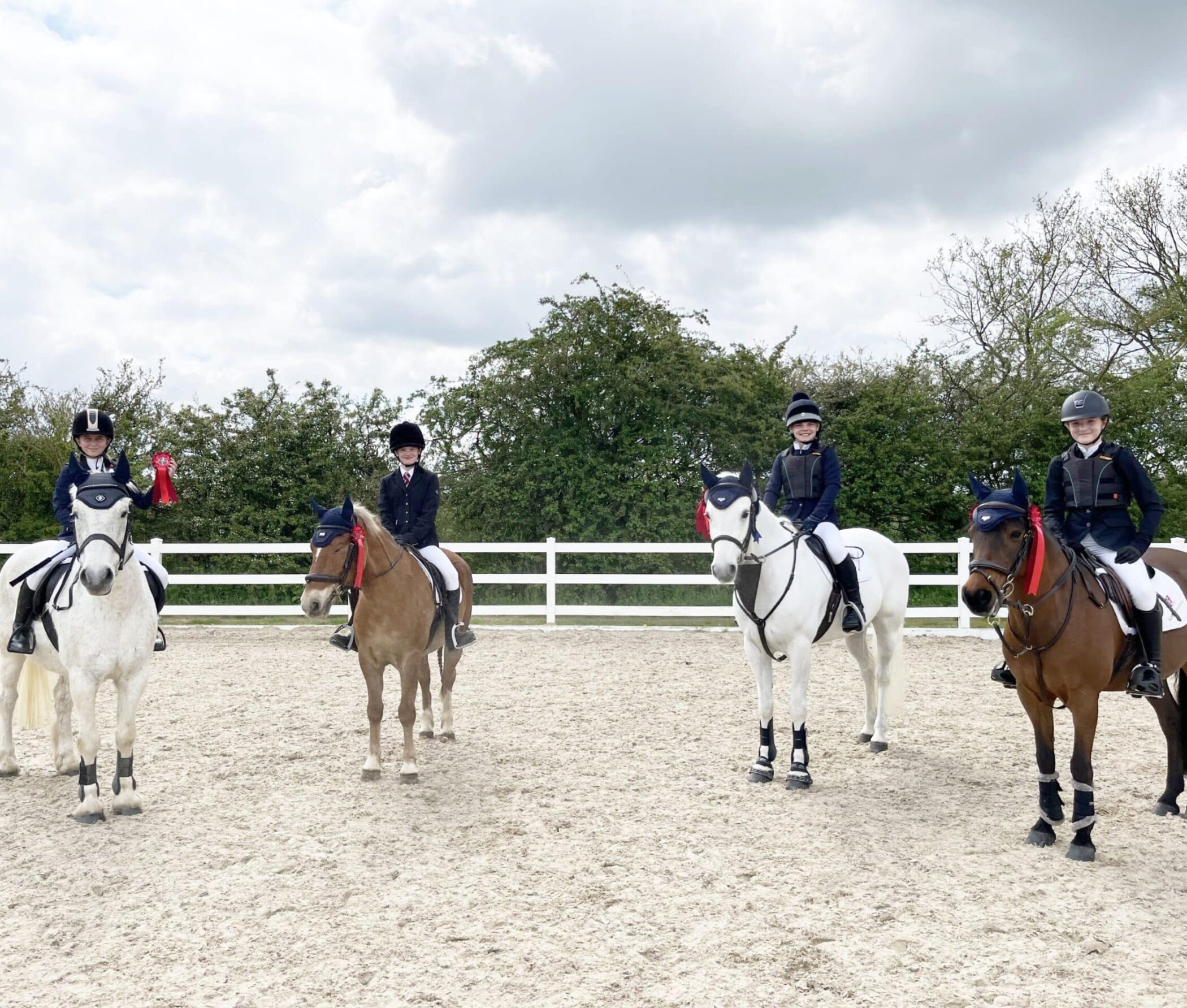 Four horse riders in riding kit sit on their horses in an outdoor arena. The rider on the left holds a red rosette. All horses wear bridles and ear bonnets. A white fence and green trees are in the background under a cloudy sky.