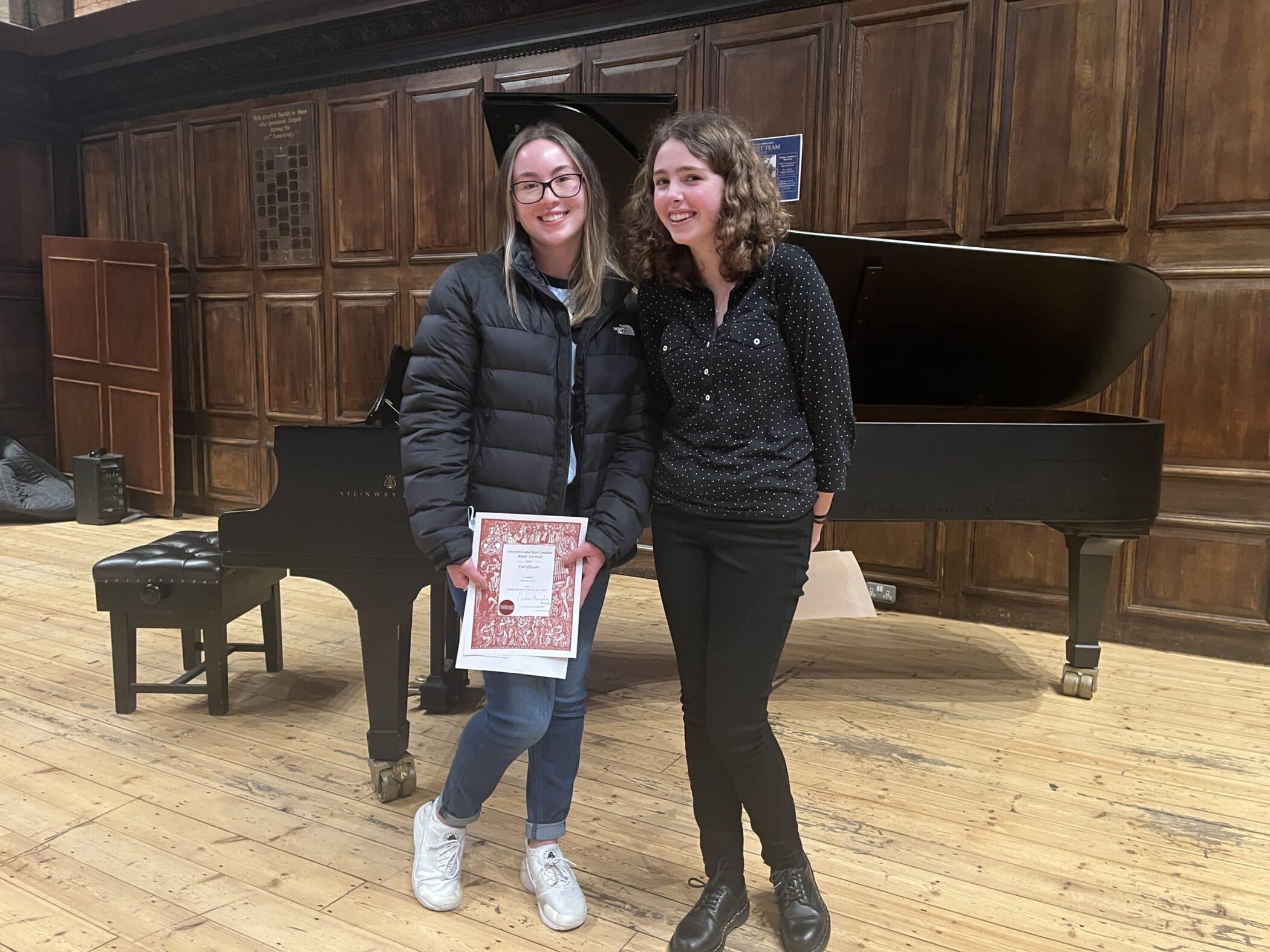 Two young women stand smiling in front of a grand piano on a wooden stage. One holds a certificate and wears glasses, a black jacket, jeans, and white trainers; the other wears a black shirt, black trousers, and black shoes.