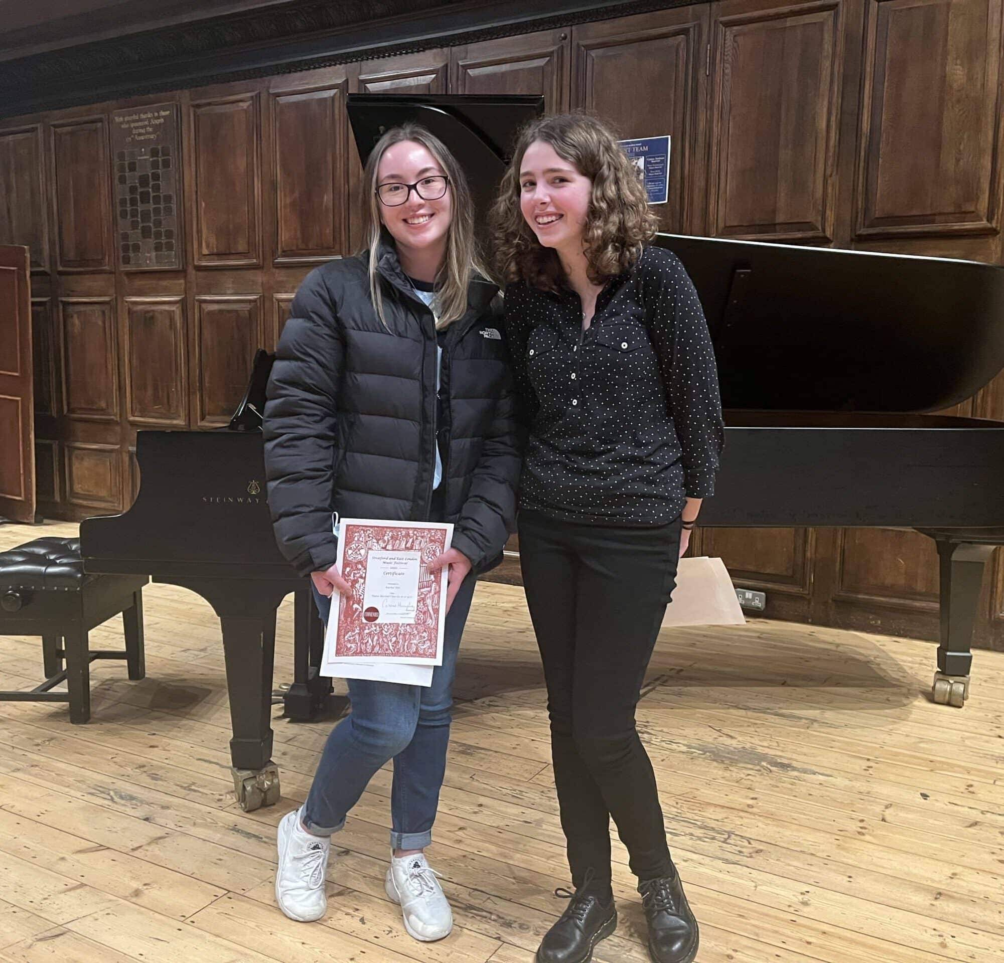 Two young women stand smiling in front of a grand piano on a wooden stage. One holds a certificate and wears glasses, a black jacket, jeans, and white trainers; the other wears a black shirt, black trousers, and black shoes.