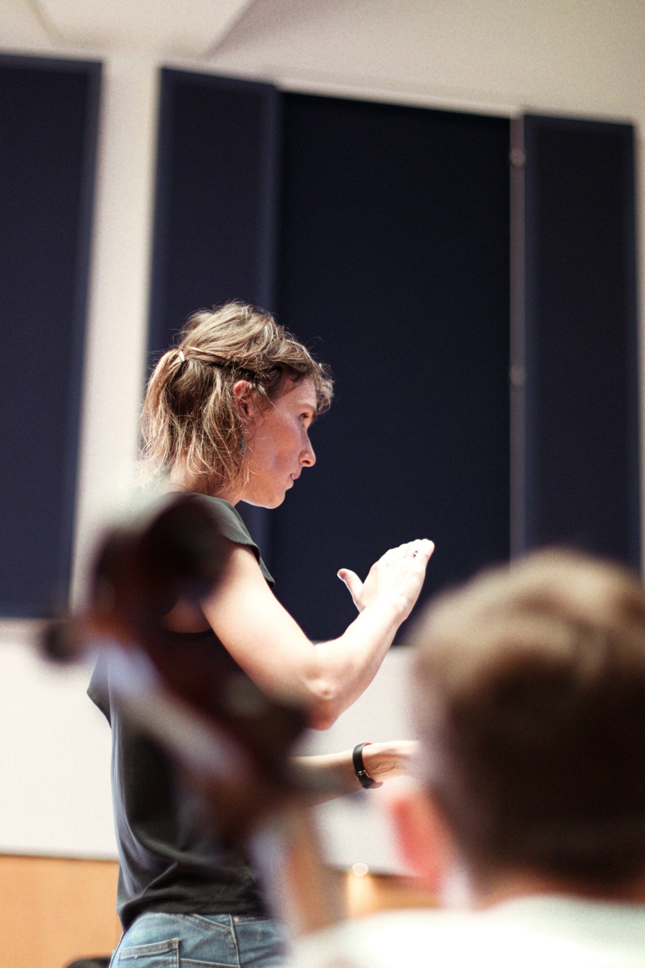 A person stands with one arm raised, appearing to conduct or give instructions, in a room with blue and white walls. The blurred foreground suggests an audience or group of musicians.