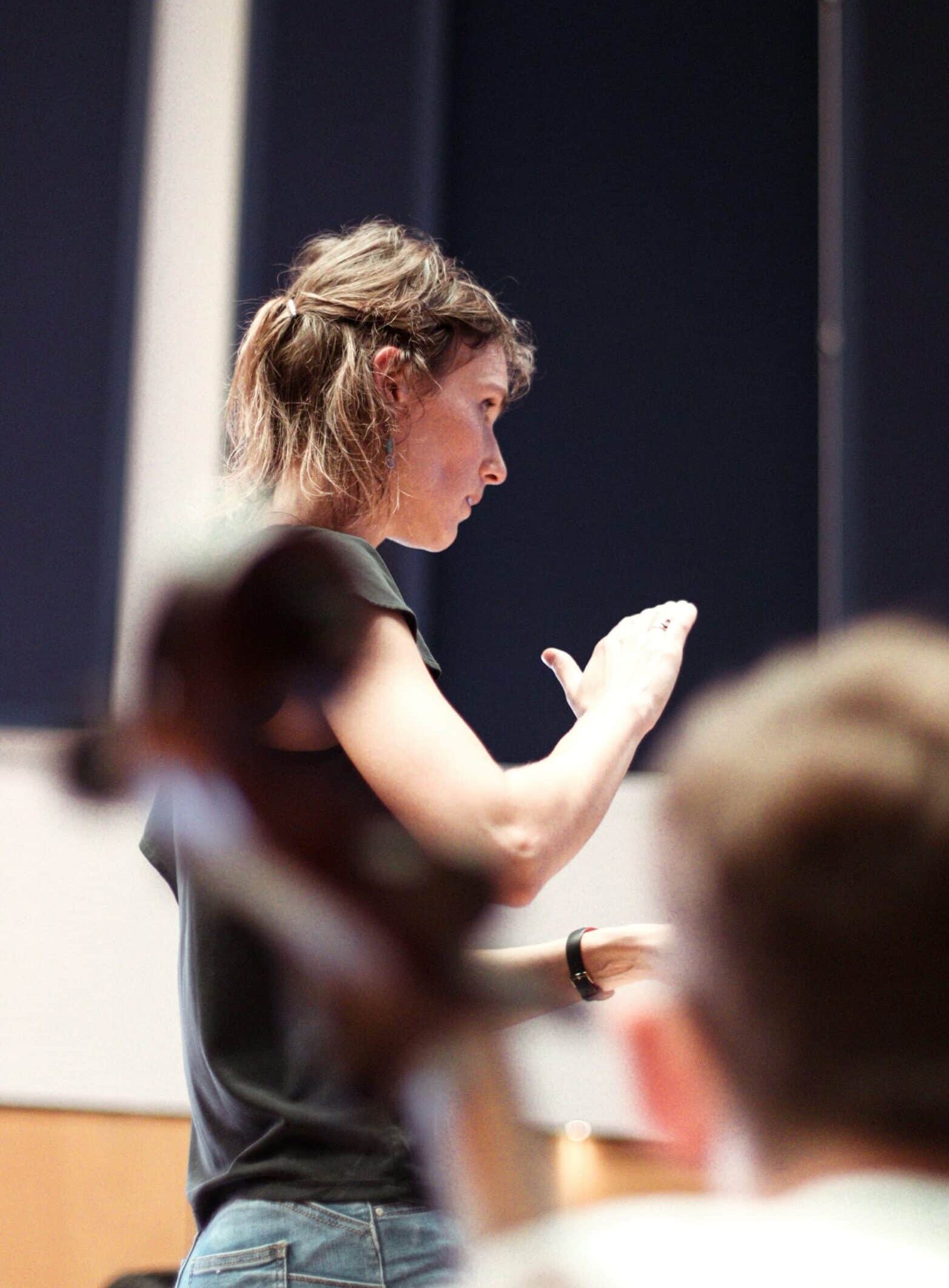 A person stands with one arm raised, appearing to conduct or give instructions, in a room with blue and white walls. The blurred foreground suggests an audience or group of musicians.