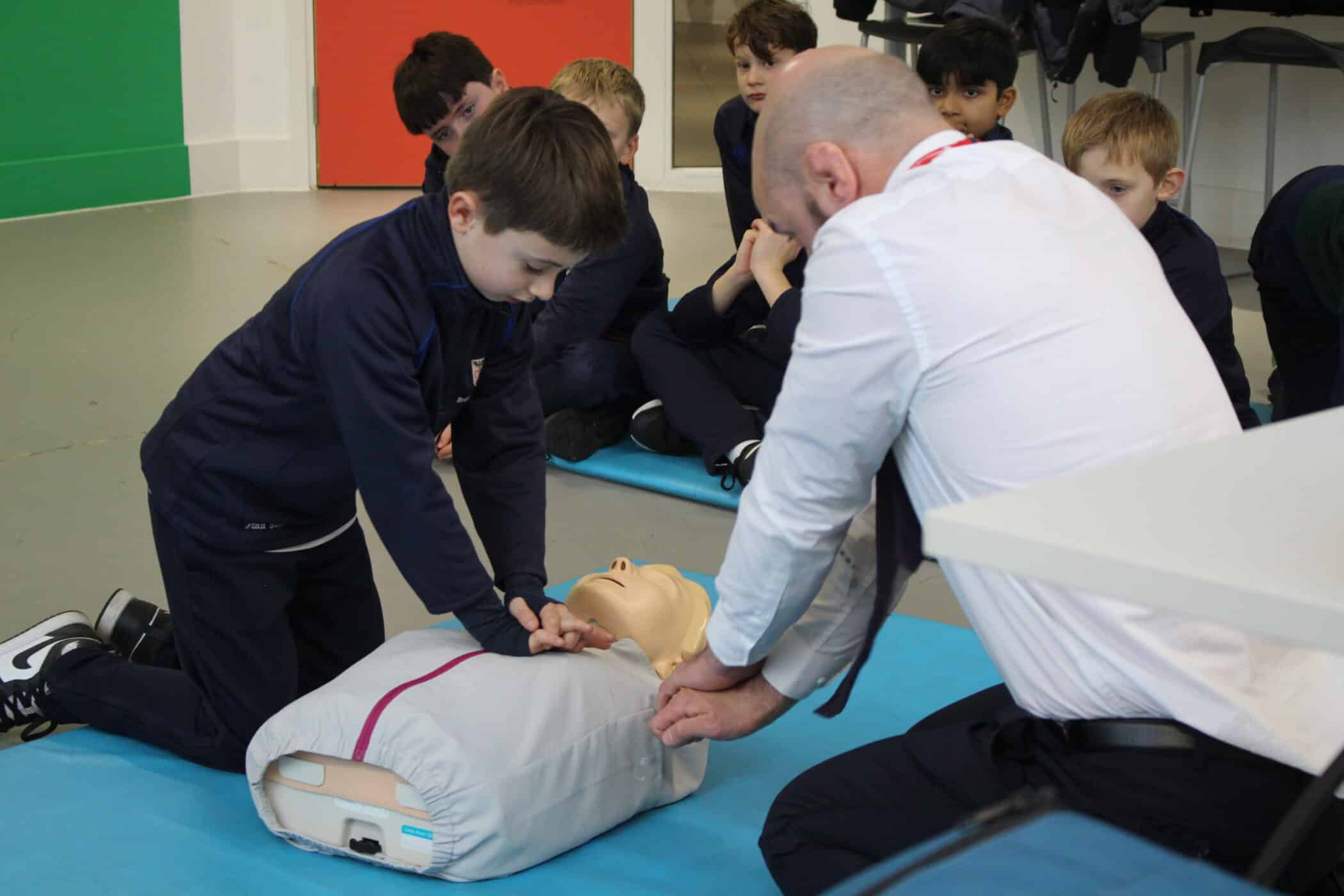 A boy practises CPR on a manikin while an instructor guides him; other children watch attentively in the background.