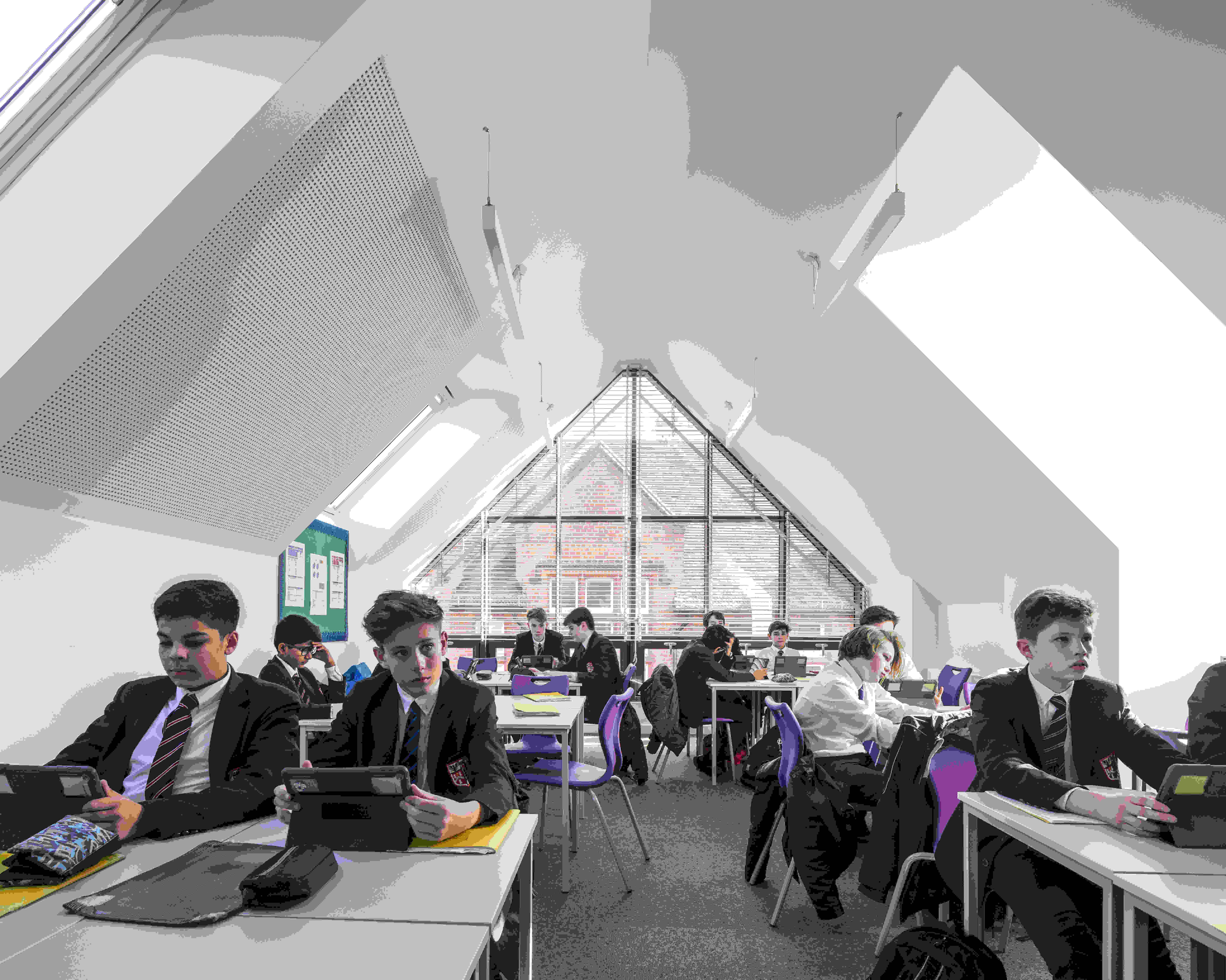 An academic classroom with students in school uniforms sits at desks, using tablets. The space features a sloped ceiling and large triangular windows that fill the room with natural light.
