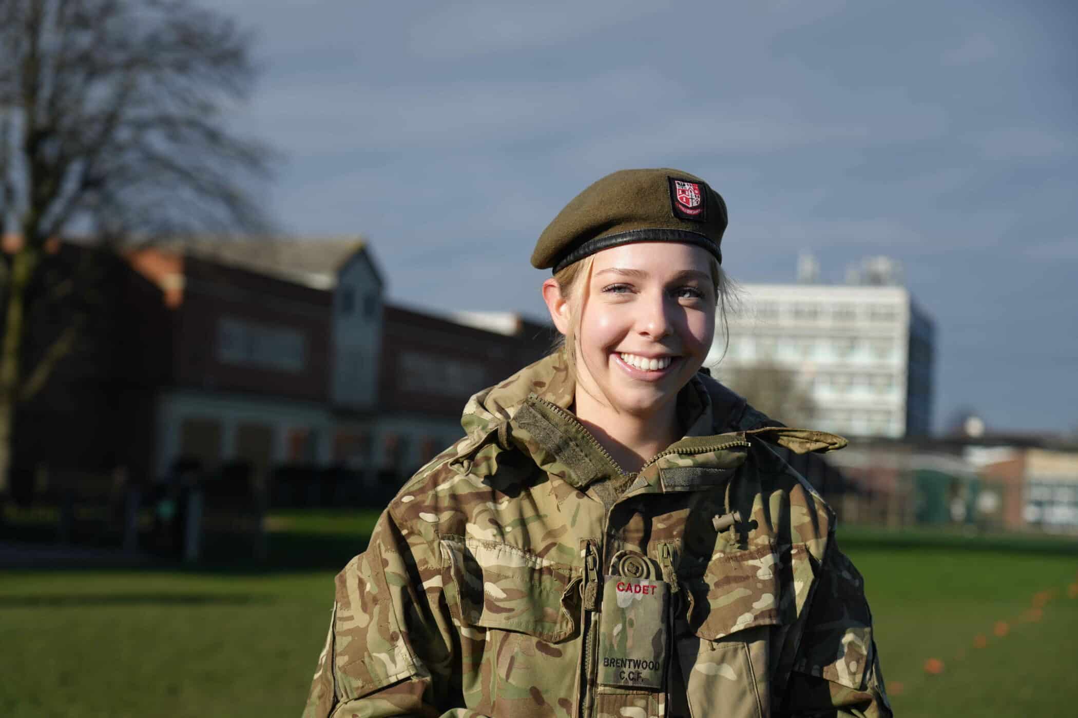 A young woman in military camouflage uniform and beret stands outdoors on a grassy field, smiling at the camera, representing her involvement in co-curricular activities, with buildings and a tree in the background under a partly cloudy sky.