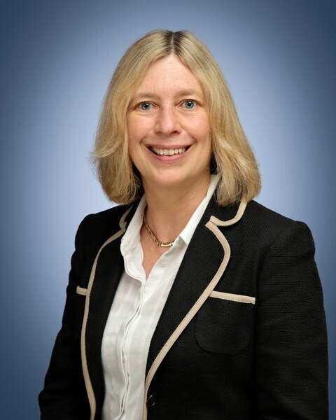 A woman with shoulder-length blonde hair, wearing a black blazer with white trim over a white blouse, smiles at the camera against a plain blue background, presenting herself as the Senior Deputy Headteacher.