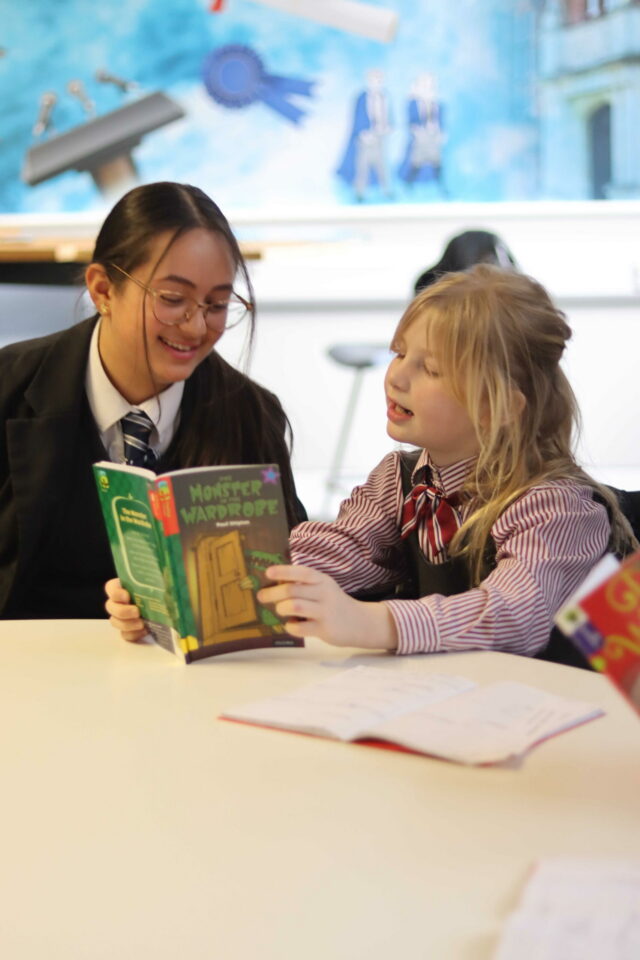 Two children in school uniforms sit at a table, smiling and reading a book titled “Monster in the Wardrobe” together. An open exercise book lies in front of them. The bright classroom background features a colourful School Shop display.
