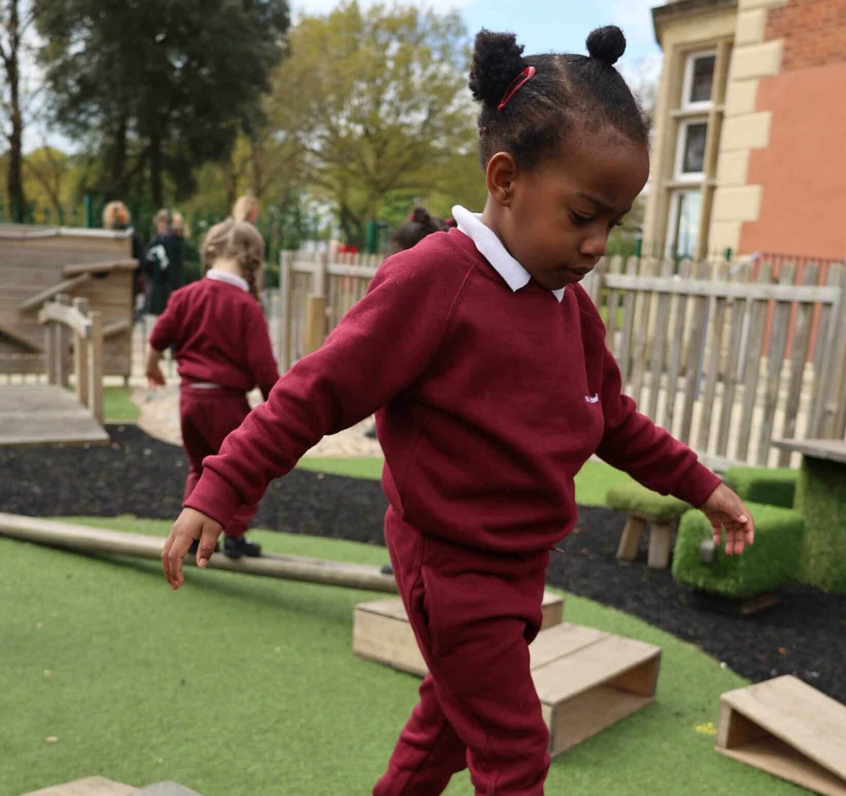 A young child in a maroon Brentwood Preparatory School uniform balances on a wooden beam in an outdoor playground, with another child in a similar uniform walking nearby. A wooden fence and building can be seen in the background.