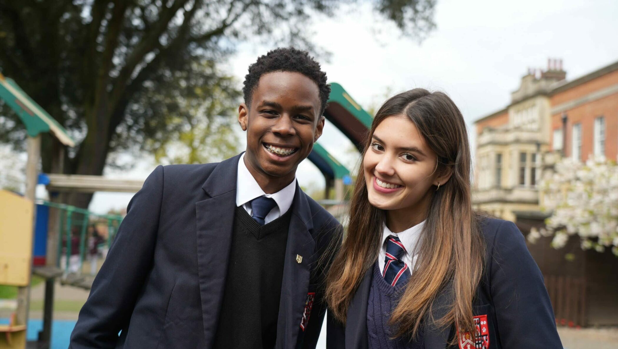 Two students in school uniforms, a boy and a girl, smile outdoors at Brentwood School Sixth Form, with the school building and playground equipment in the background.