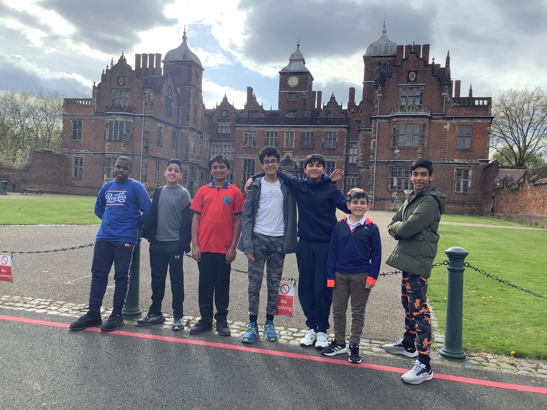 Seven boys stand in a row smiling in front of an old, large, red-brick building with towers and chimneys on a cloudy day. A No Parking sign is visible on the ground in front of them.