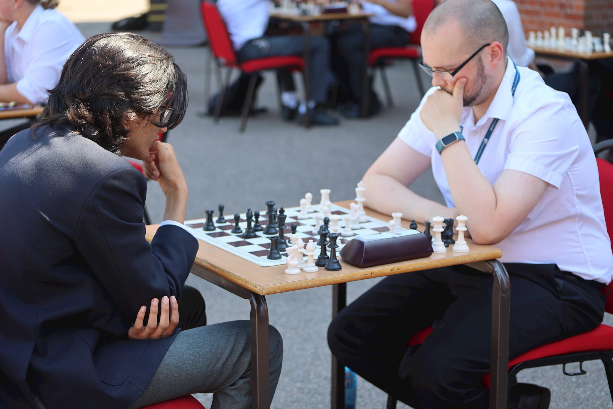 A man in a white shirt and glasses sits at a table outdoors, deep in thought whilst playing a game of chess. The chessboard is set up with pieces, and other people are seen playing chess in the background.