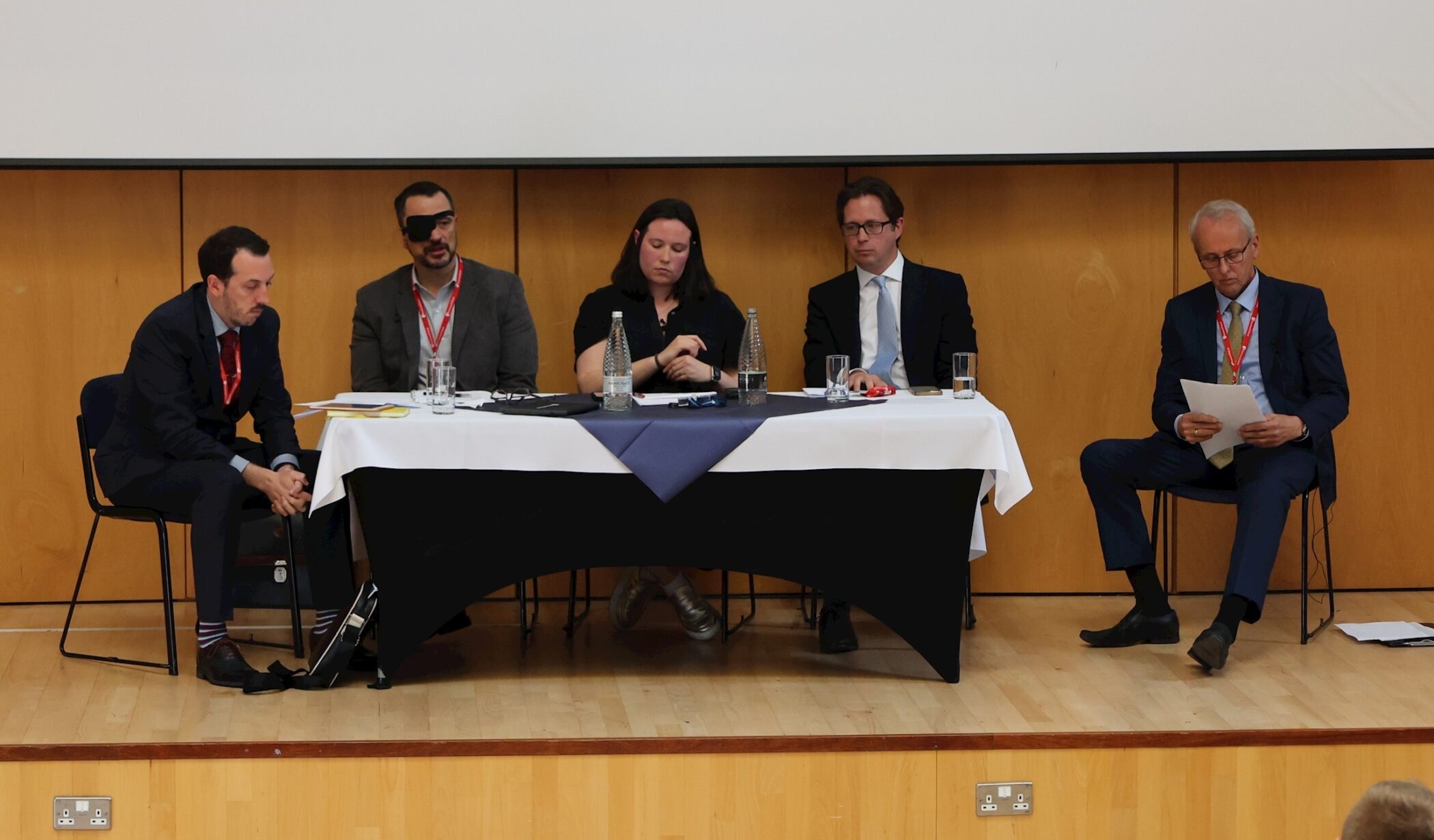 Five people in formal attire sit at a table on a stage with water bottles and papers, appearing to prepare for or participate in a panel discussion in a wood-panelled room.