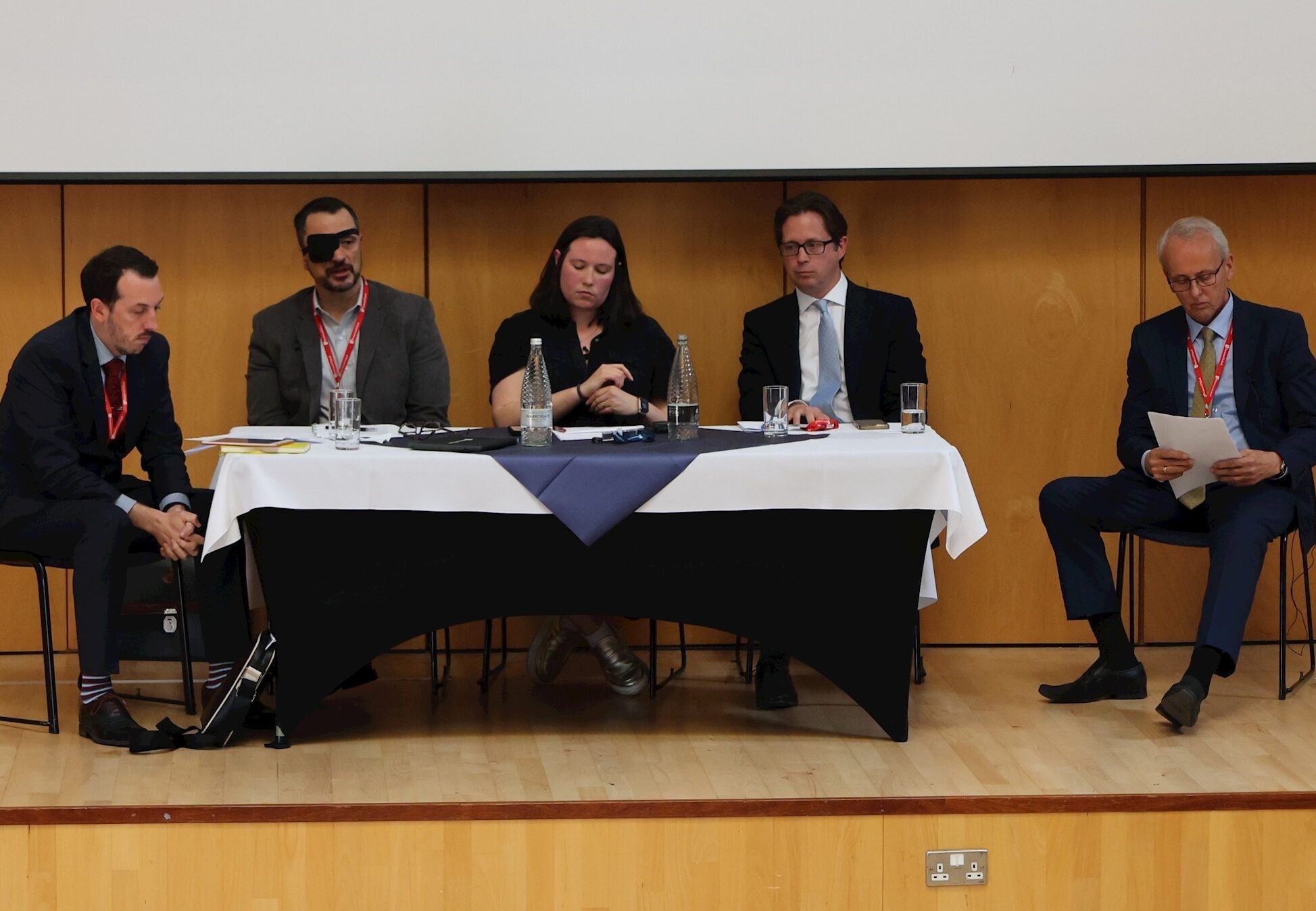 Five people in formal attire sit at a table on a stage with water bottles and papers, appearing to prepare for or participate in a panel discussion in a wood-panelled room.