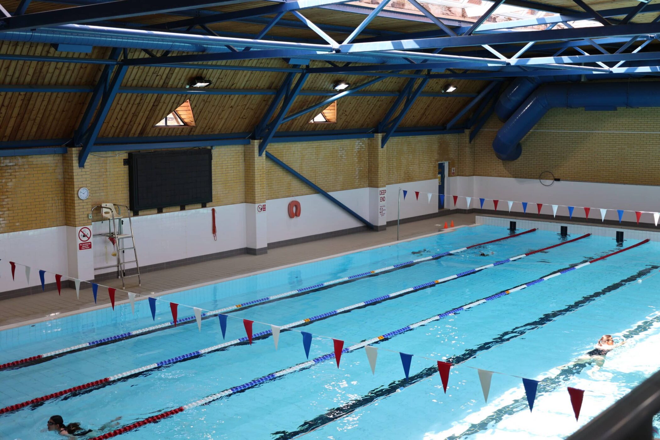 The indoor swimming pool at Brentwood School Sports Centre features several lanes marked by red, blue, and white dividers. A few swimmers are in the water, surrounded by yellow brick walls, a lifeguard’s chair, and a high blue ceiling with exposed beams.