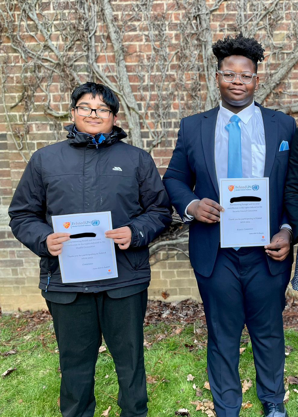 Two young men stand side by side outdoors, smiling and holding certificates. One wears glasses and a black jacket, while the other is dressed in a navy suit with a blue tie. They are in front of a brick wall with vines.