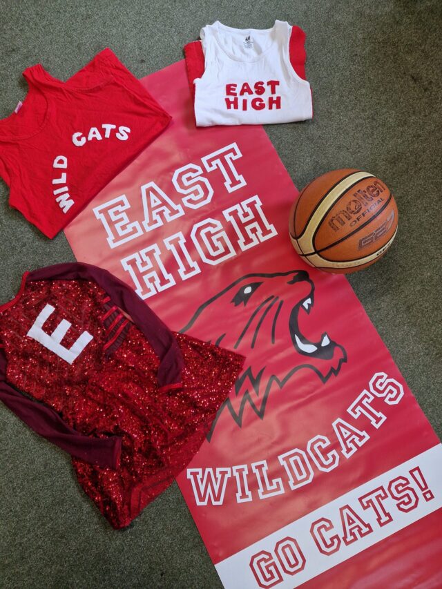 Three red and white East High Wildcats shirts, a red cheerleading outfit, a basketball, and a large East High Wildcats Go Cats! banner with a wildcat logo are arranged on a grey carpet.