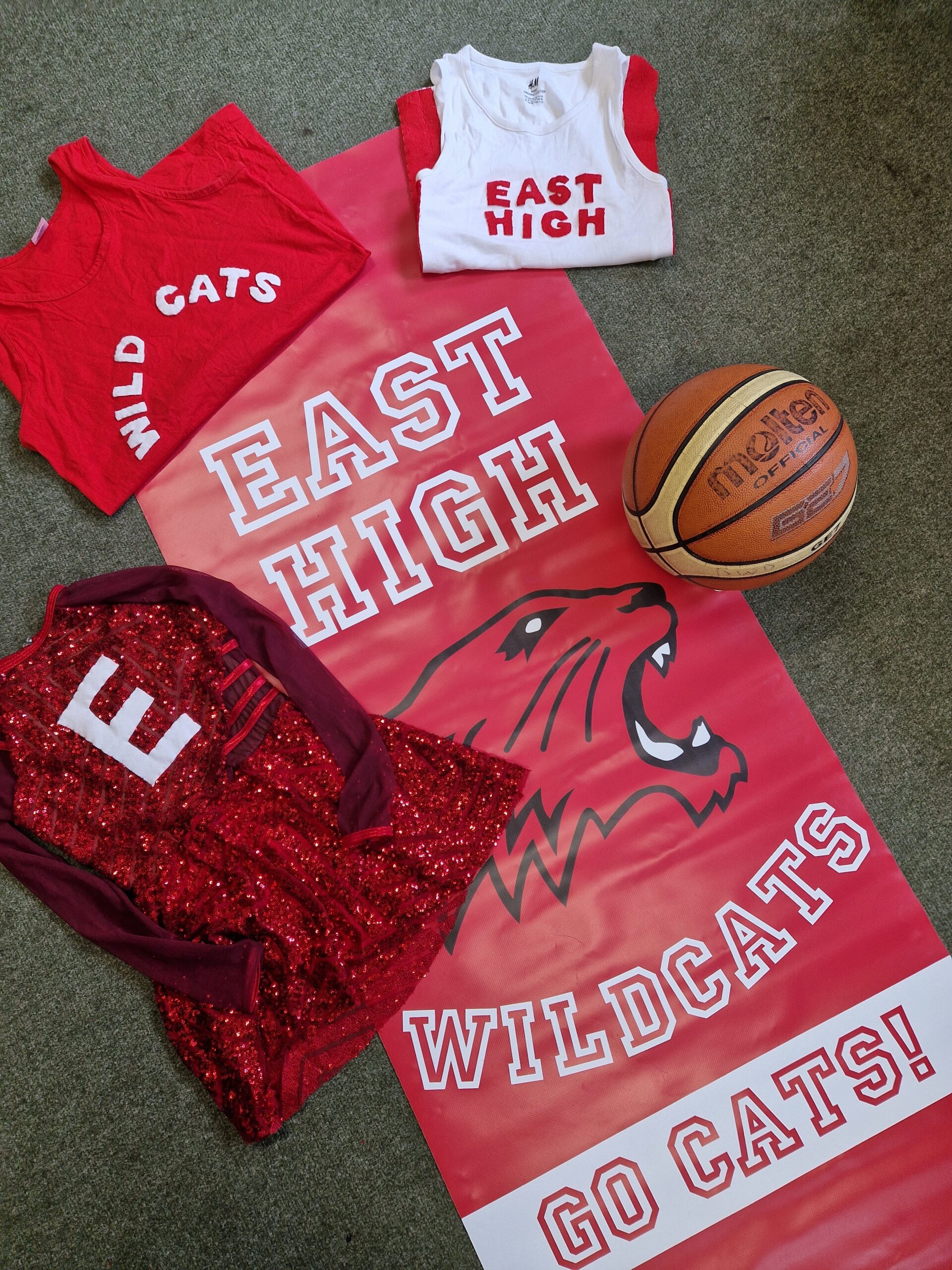 Three red and white East High Wildcats shirts, a red cheerleading outfit, a basketball, and a large East High Wildcats Go Cats! banner with a wildcat logo are arranged on a grey carpet.