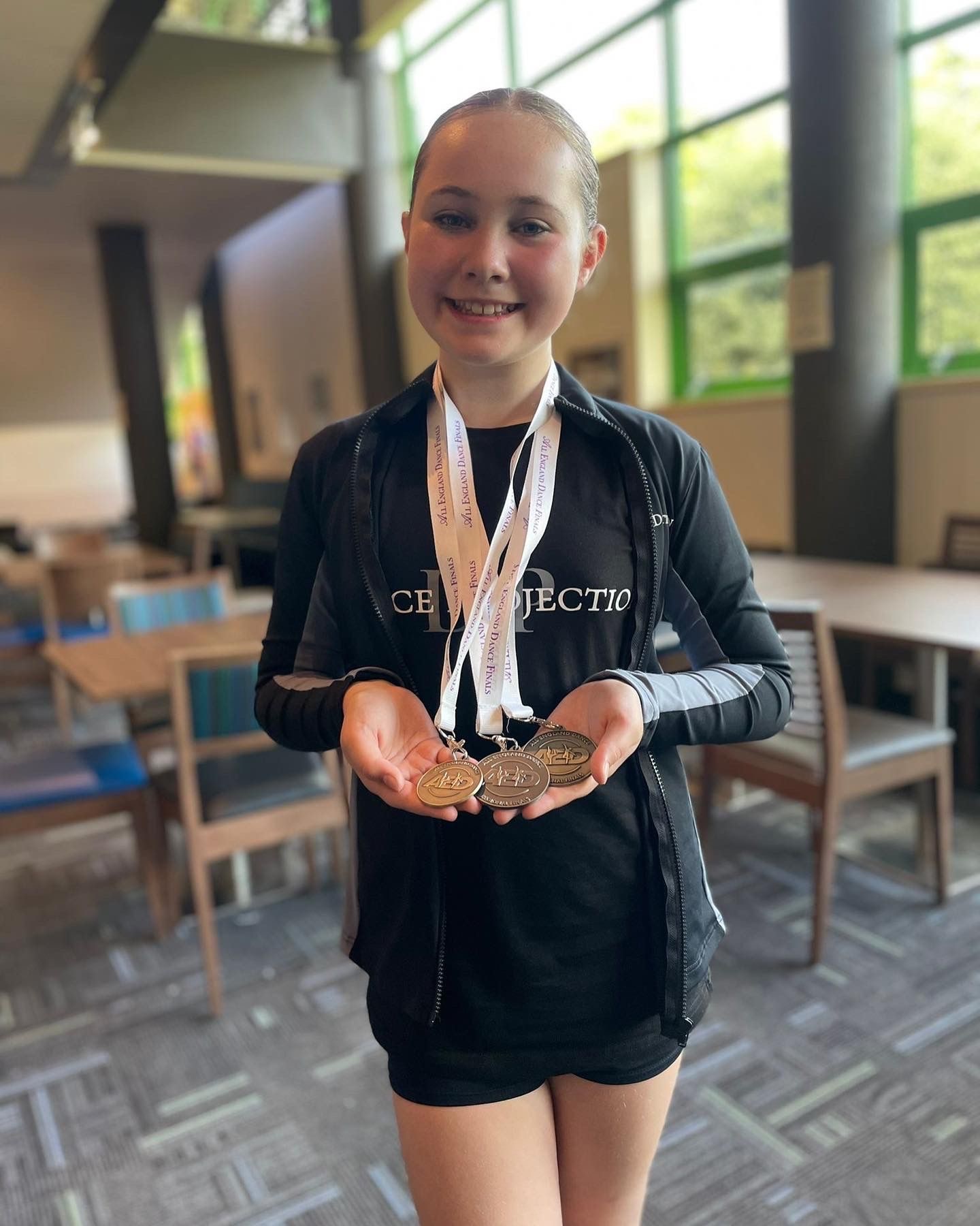 A smiling girl in sportswear stands indoors, holding three medals with white ribbons around her neck. The background shows tables, chairs, and large windows letting in natural light.