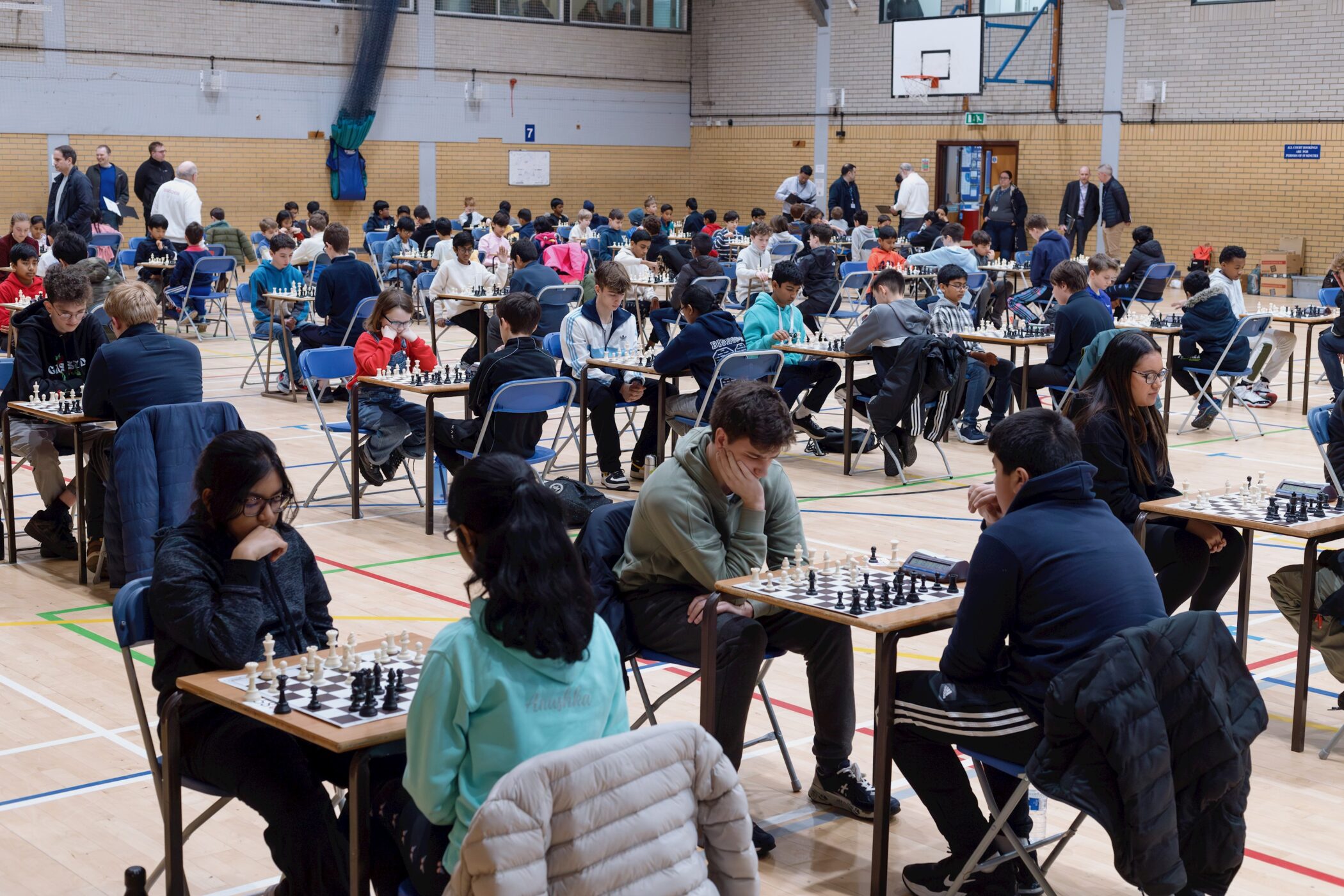 A large group of people, mostly teenagers, sit in pairs at tables playing chess in a gymnasium. The scene is busy, with many focused on their games and some standing around observing.