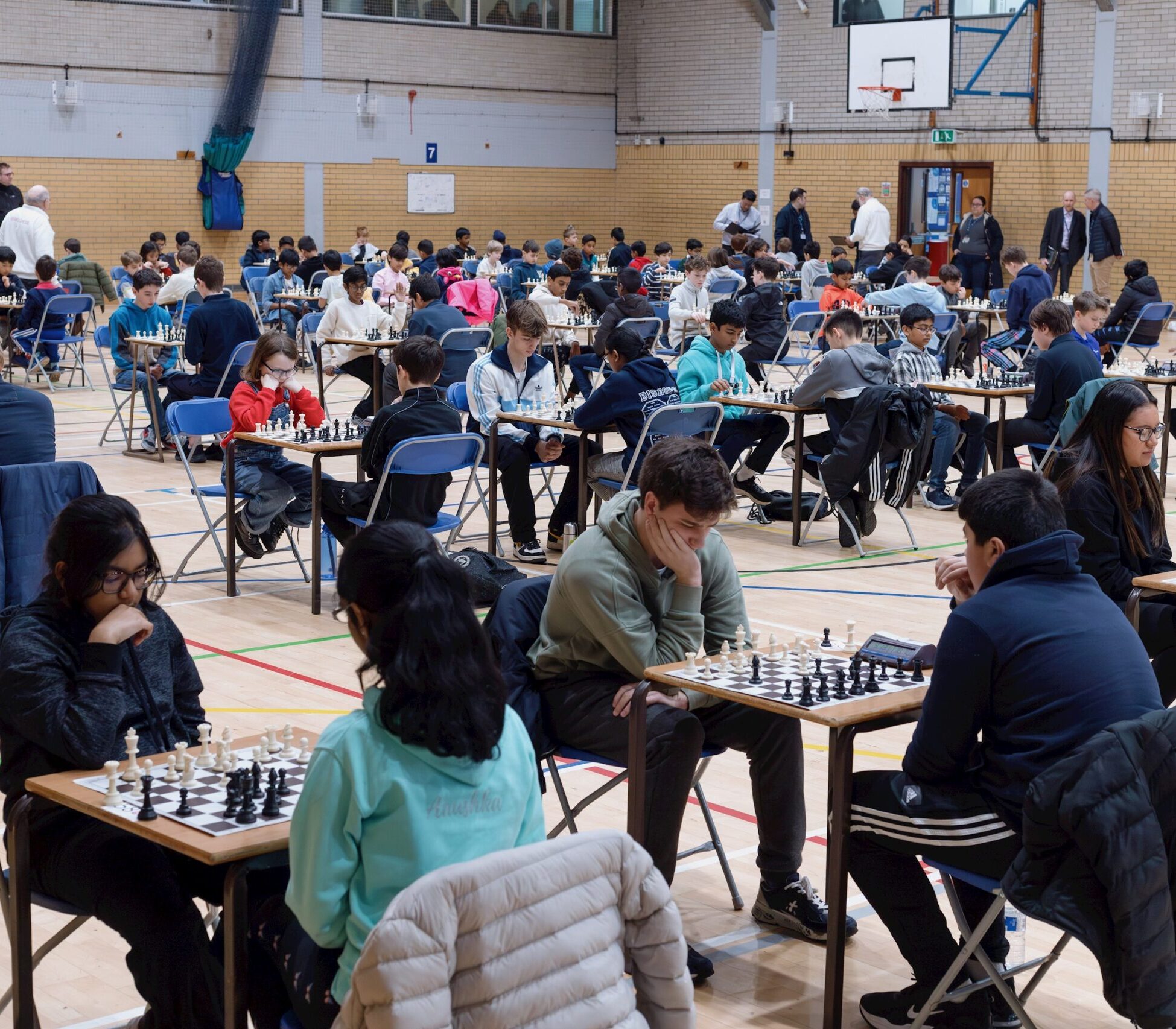 A large group of people, mostly teenagers, sit in pairs at tables playing chess in a gymnasium. The scene is busy, with many focused on their games and some standing around observing.