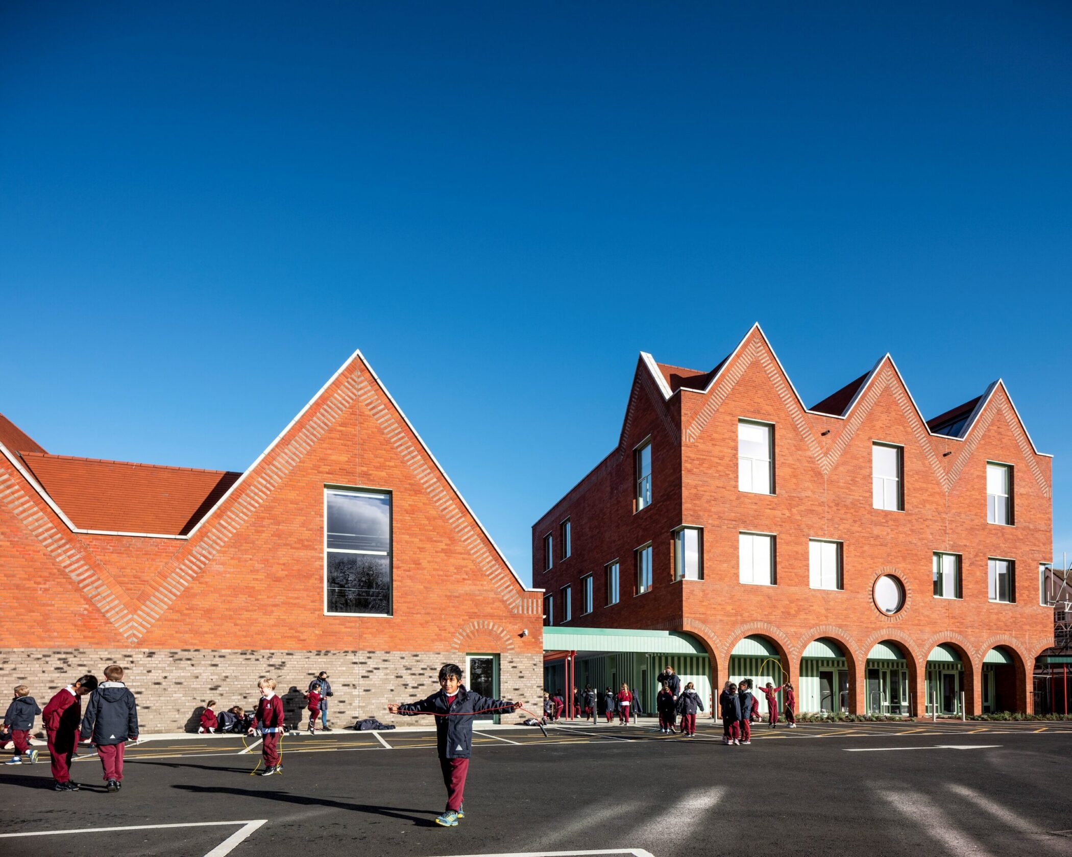 Children in uniforms play outside near modern red-brick school buildings with steep gabled roofs, large windows, and an arched facade, under a clear blue sky.