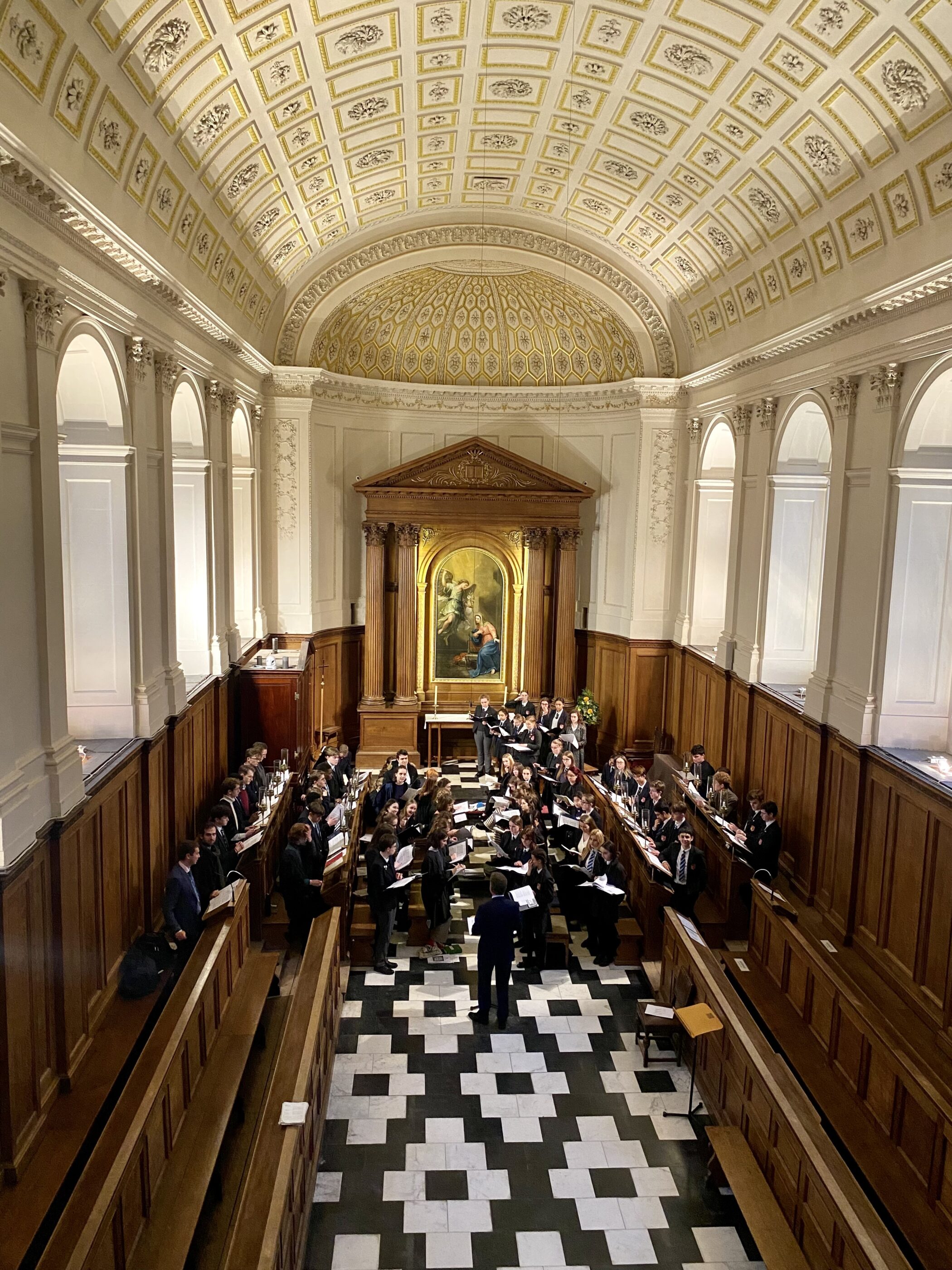 A choir sings in a grand, ornate hall with arched windows, tall wood panelling, and a decorated vaulted ceiling. A conductor stands in front, and a large painting is displayed at the back of the room.