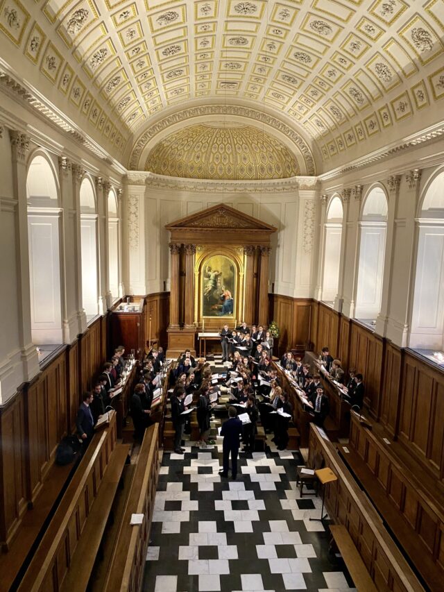 A choir sings in a grand, ornate hall with arched windows, tall wood panelling, and a decorated vaulted ceiling. A conductor stands in front, and a large painting is displayed at the back of the room.