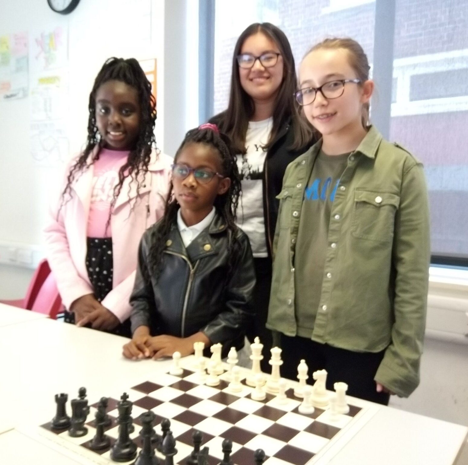 Four girls stand behind a chessboard set up for play in a classroom, smiling at the camera. Drawings and papers are visible on the white wall behind them.