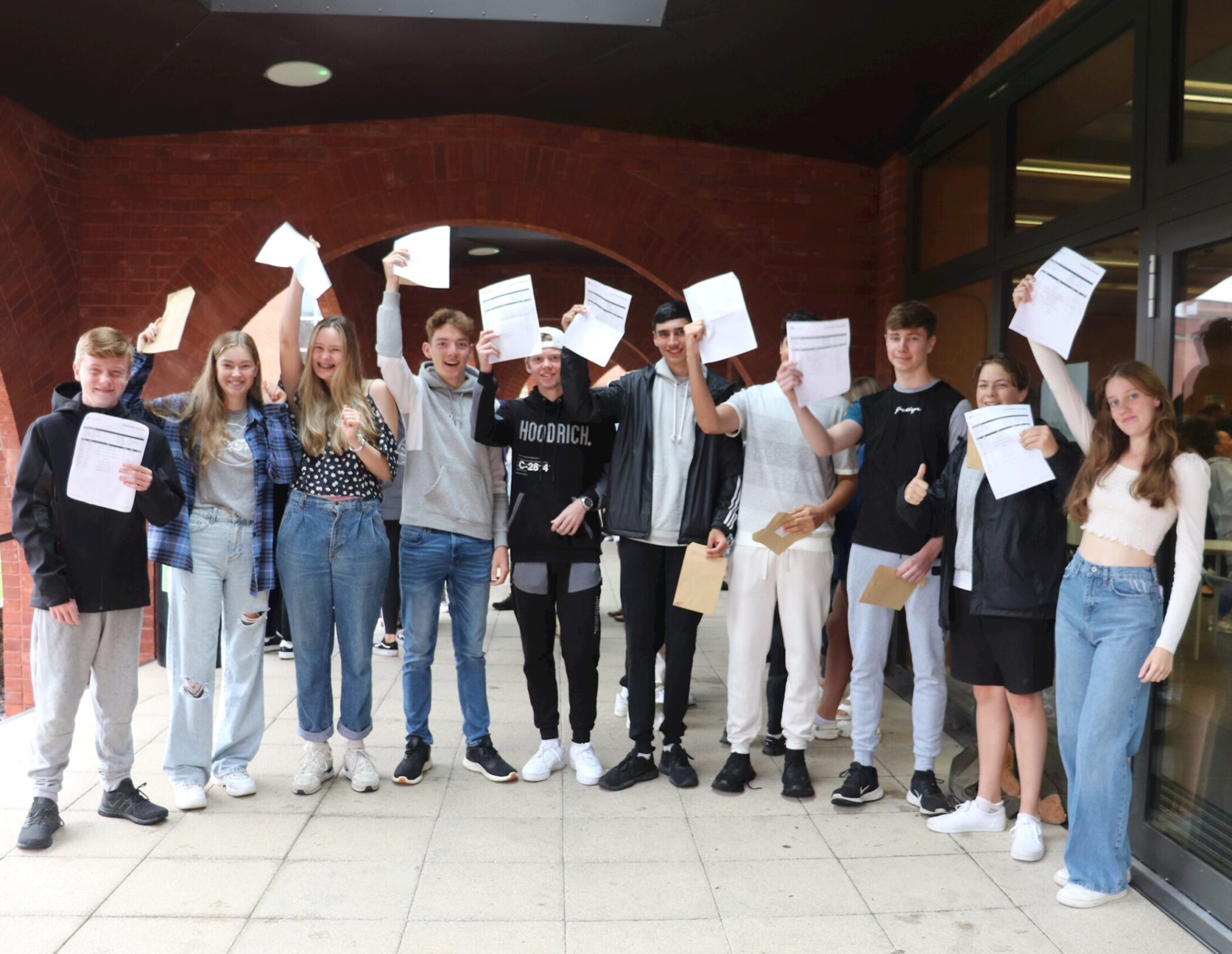 A group of smiling teenagers stand outside, holding up pieces of paper, celebrating together. They appear happy and excited, possibly after receiving exam results.