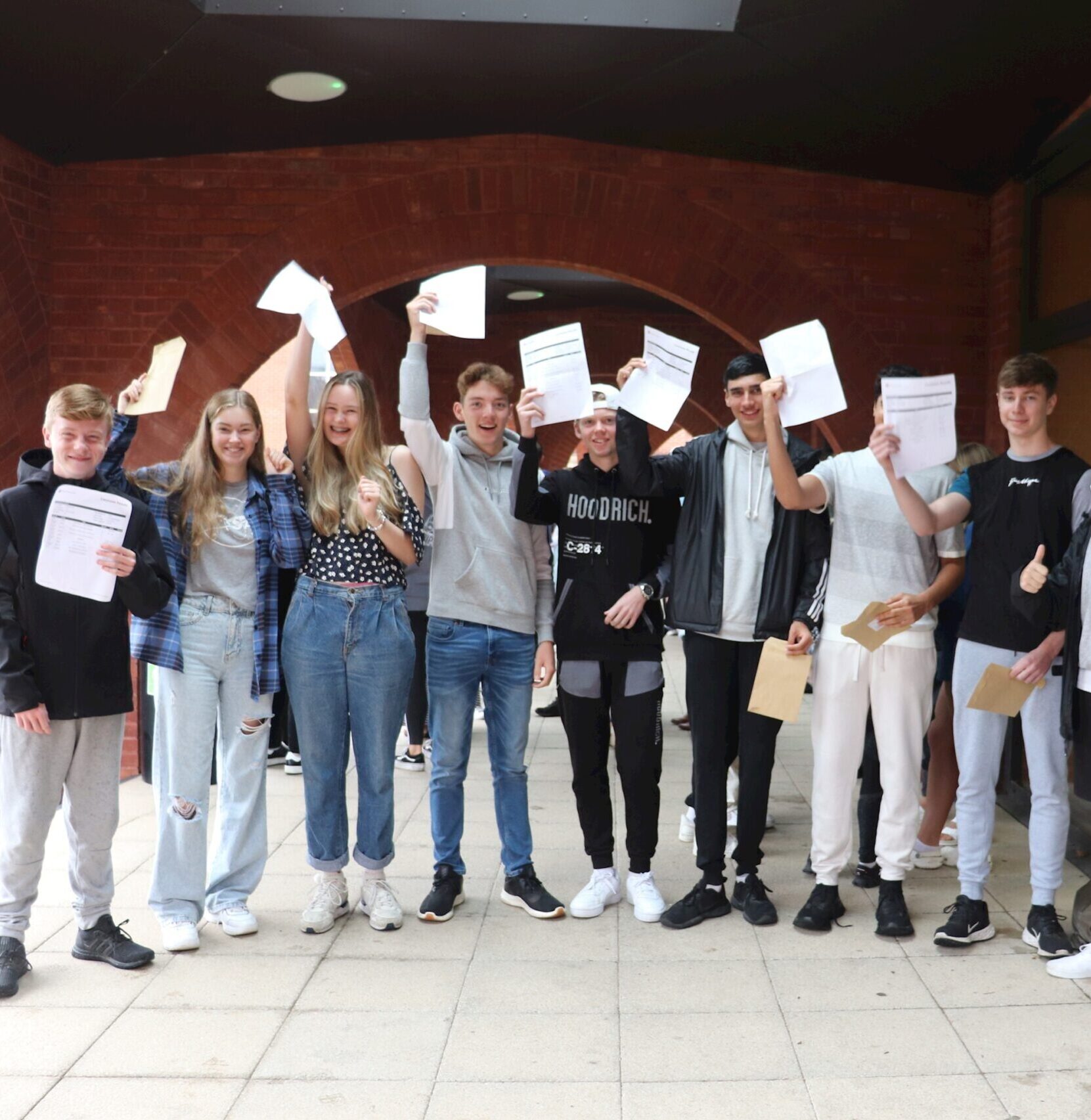 A group of smiling teenagers stand outside, holding up pieces of paper, celebrating together. They appear happy and excited, possibly after receiving exam results.