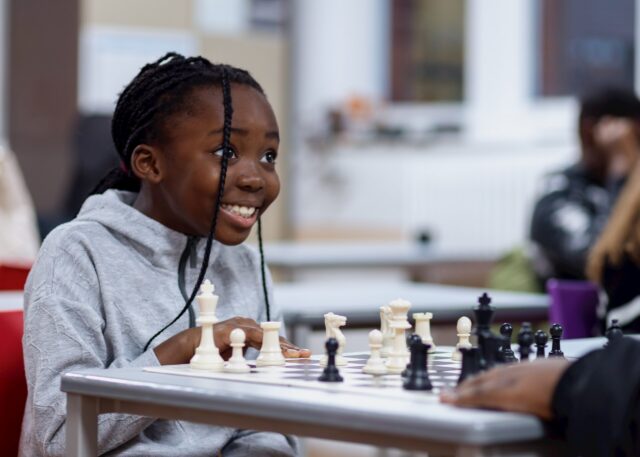 A smiling young girl with plaited hair sits at a table, playing chess with an opponent. She looks excited and engaged, wearing a grey hoodie in a bright indoor setting.