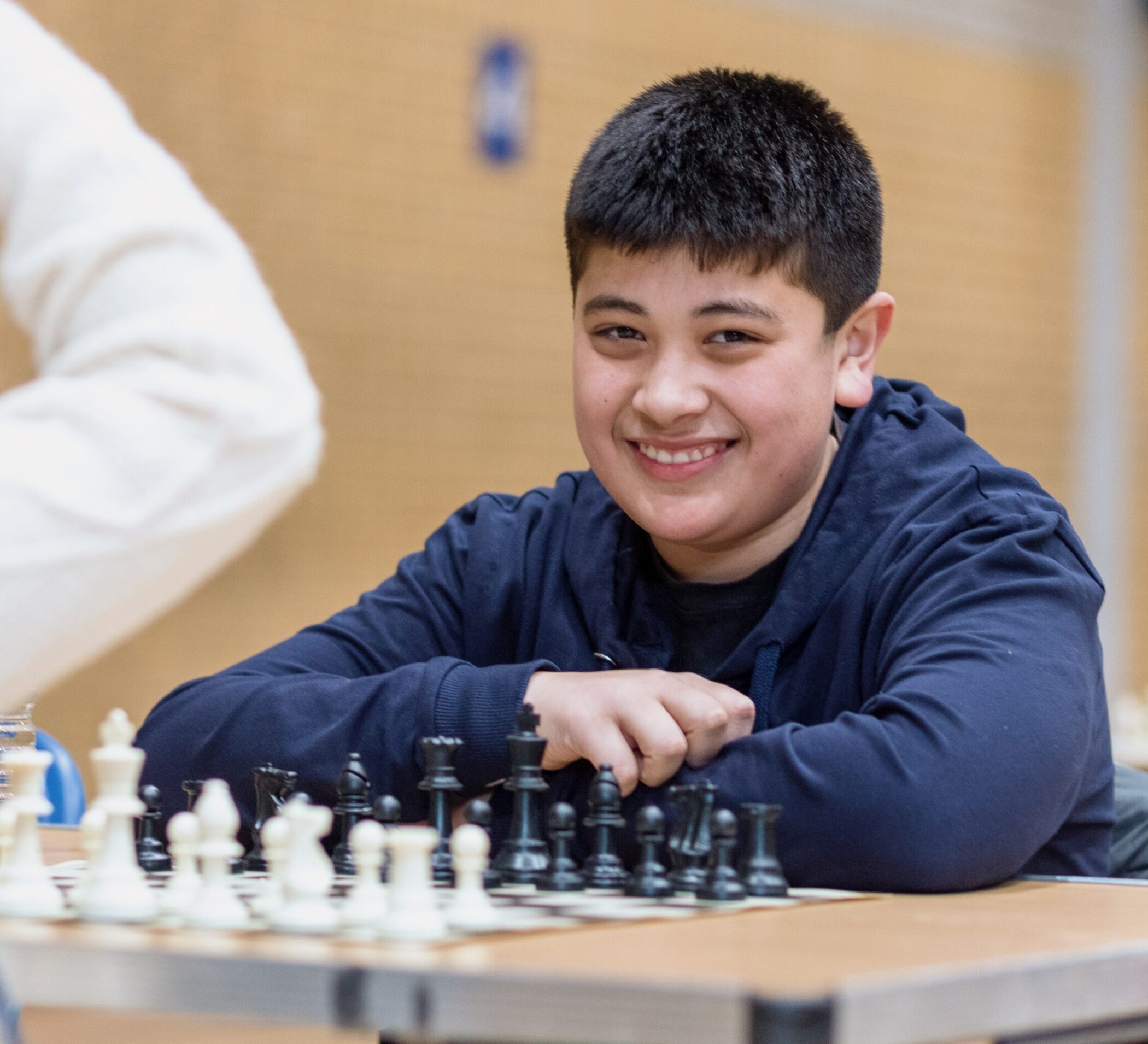 A smiling boy sits at a table, ready to play chess. He is wearing a dark hoodie and has short black hair. A chessboard with arranged pieces is in front of him.