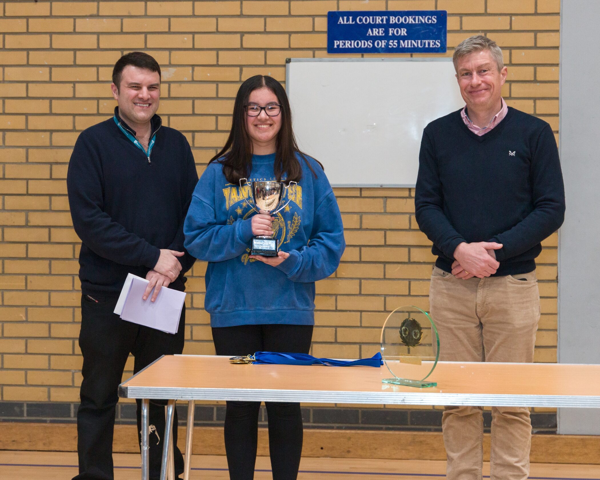 Three people stand indoors in front of a brick wall; the person in the centre holds a trophy and smiles. Two men stand on either side, one holding papers. Awards are displayed on the table in front of them.