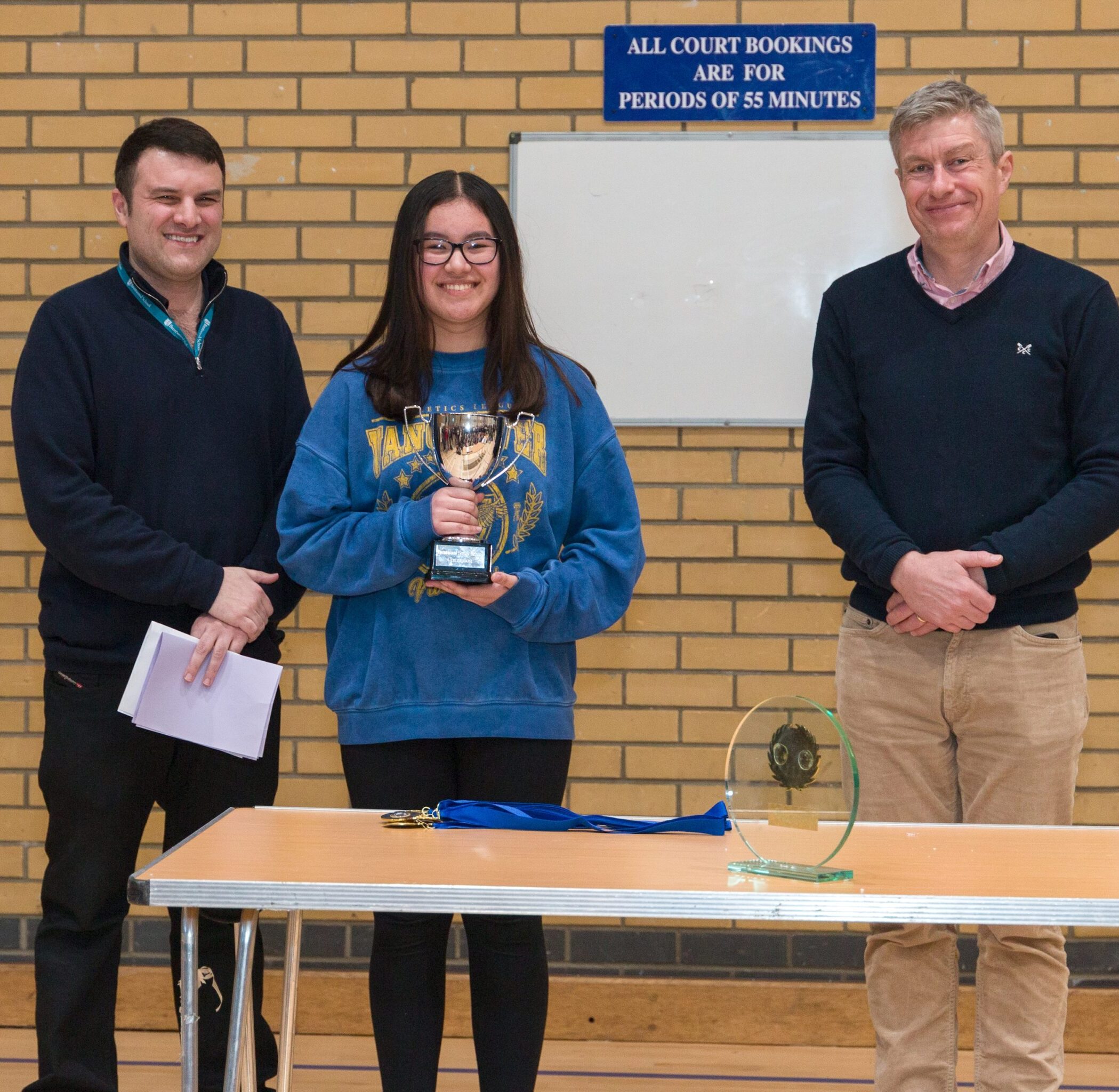Three people stand indoors in front of a brick wall; the person in the centre holds a trophy and smiles. Two men stand on either side, one holding papers. Awards are displayed on the table in front of them.