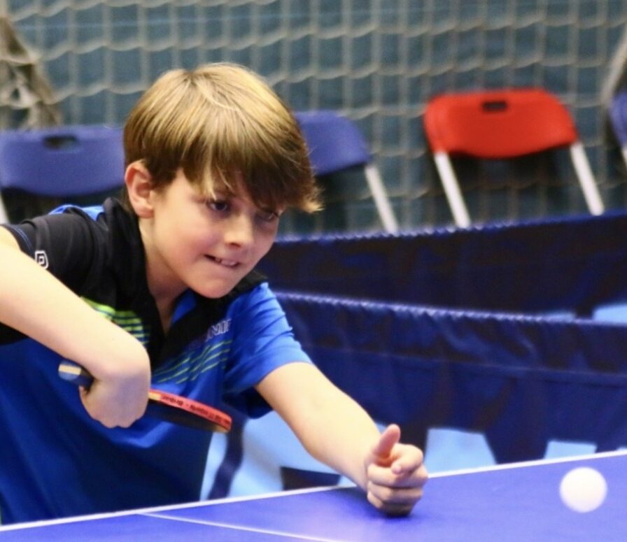 A young boy in a blue shirt concentrating as he plays table tennis, holding a bat and getting ready to hit a white table tennis ball. Blue and red chairs and a net are visible in the background.