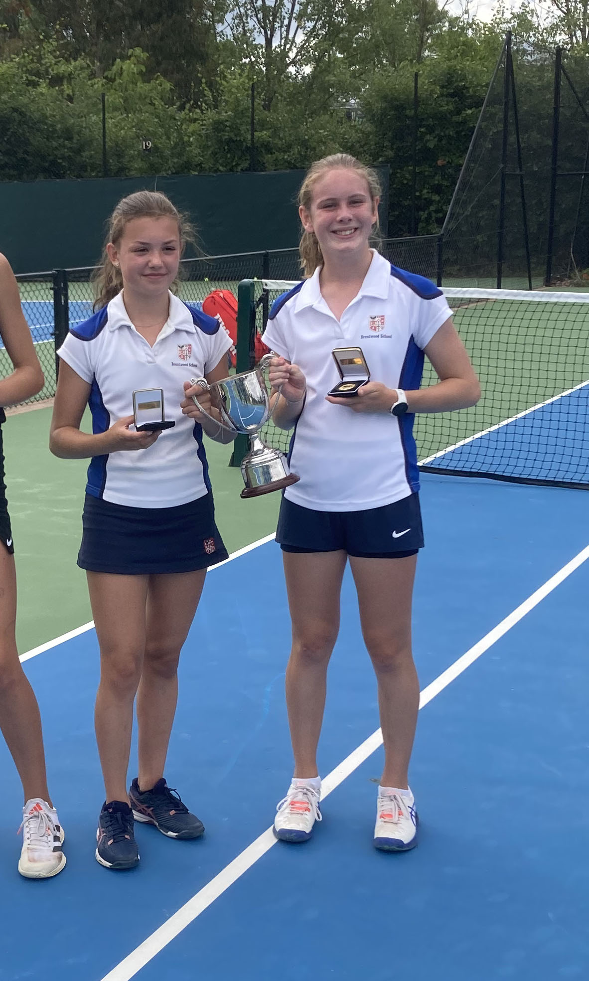 Two teenage girls in tennis kit stand on a tennis court, smiling and holding medals and a trophy. A tennis net and green trees are visible in the background.