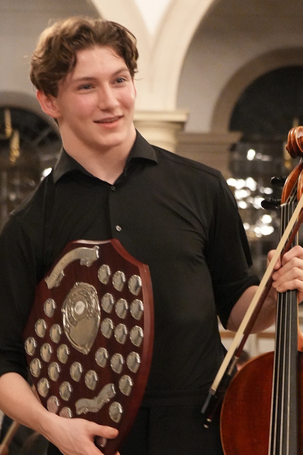 A young man in a black shirt smiles whilst holding a large shield-shaped trophy and a cello, standing indoors with arched windows in the background.