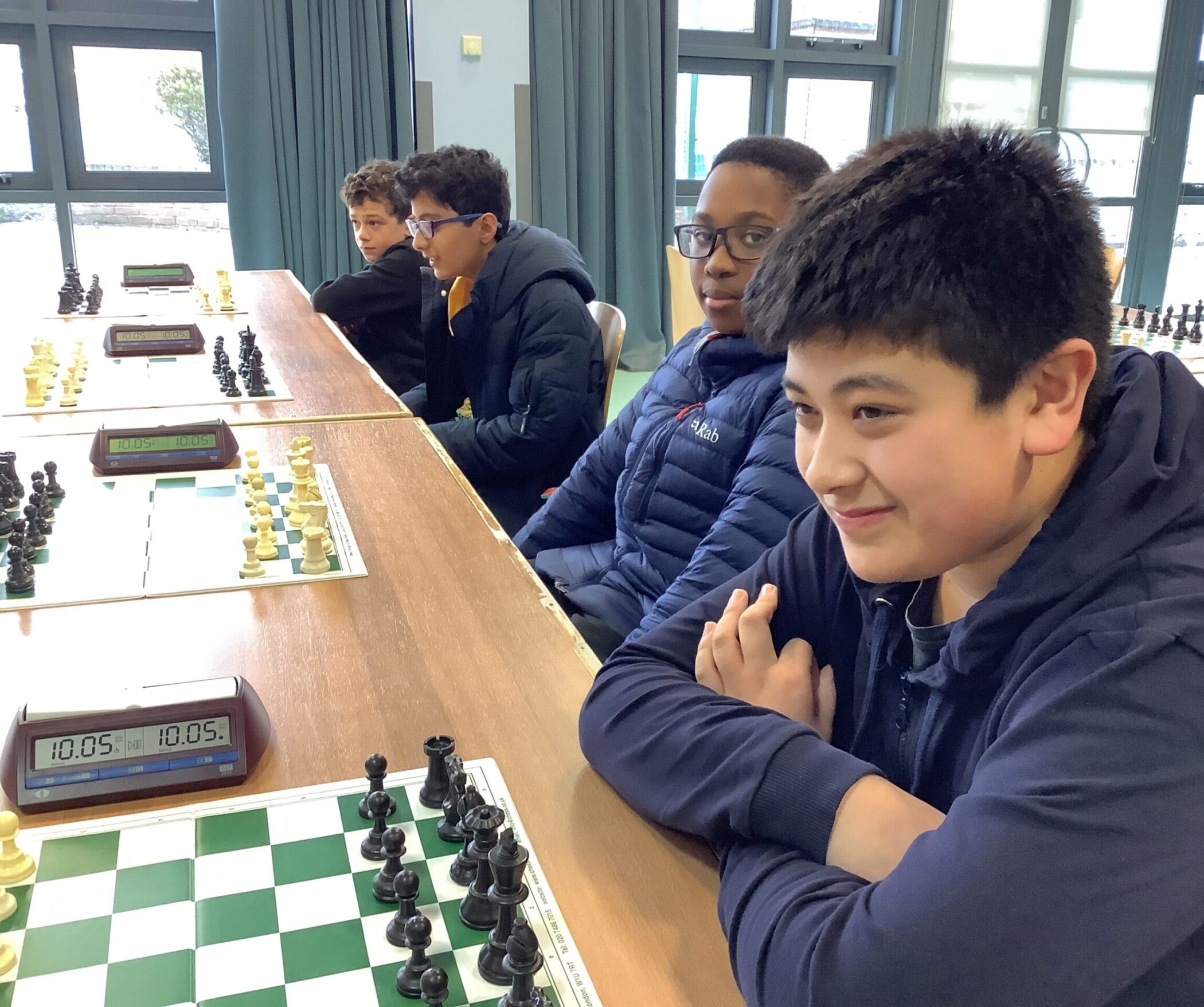 Four boys sit at a table with chess boards and timers, appearing focused and ready to play chess in a bright room with large windows.