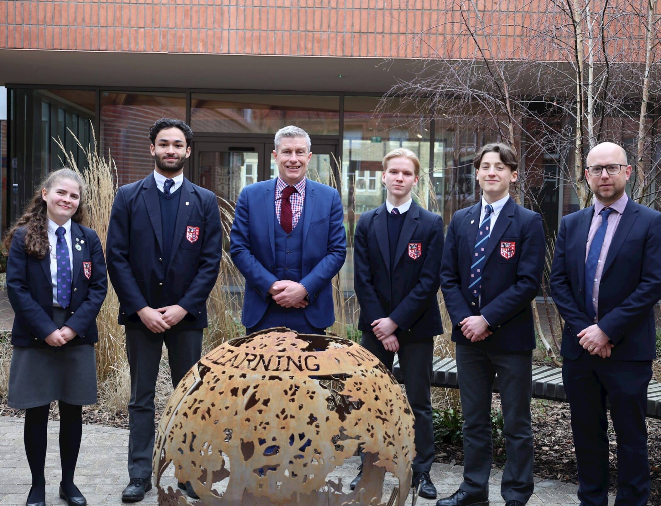 A group of five students in school uniforms and two adults in suits stand outside near a decorative metal globe sculpture with the word LEARNING on it, posing for a photo in front of a building.