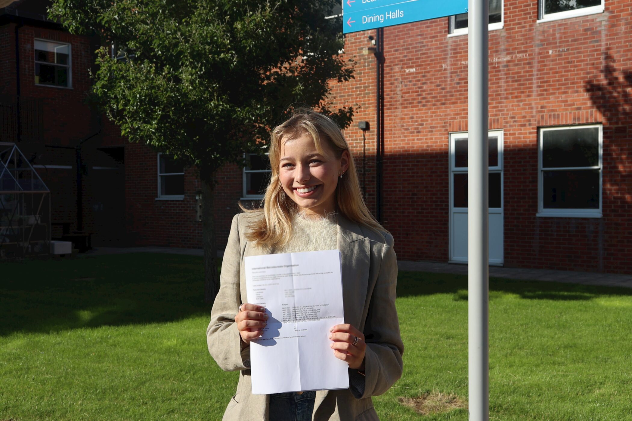 A young woman stands outside on a sunny day, smiling and holding up a printed document. She is in front of a brick building and a blue sign pointing to Dining Halls. Green grass and a tree are visible in the background.