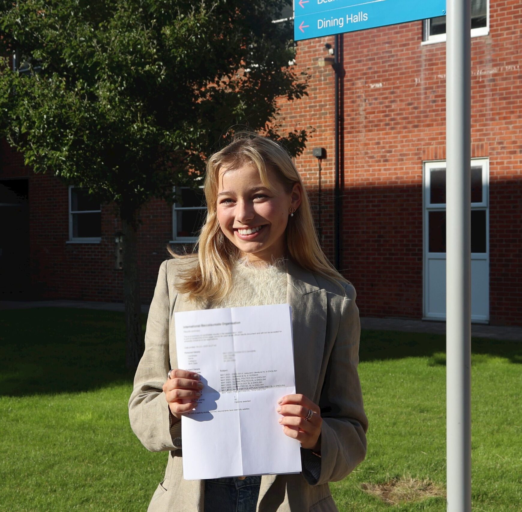 A young woman stands outside on a sunny day, smiling and holding up a printed document. She is in front of a brick building and a blue sign pointing to Dining Halls. Green grass and a tree are visible in the background.
