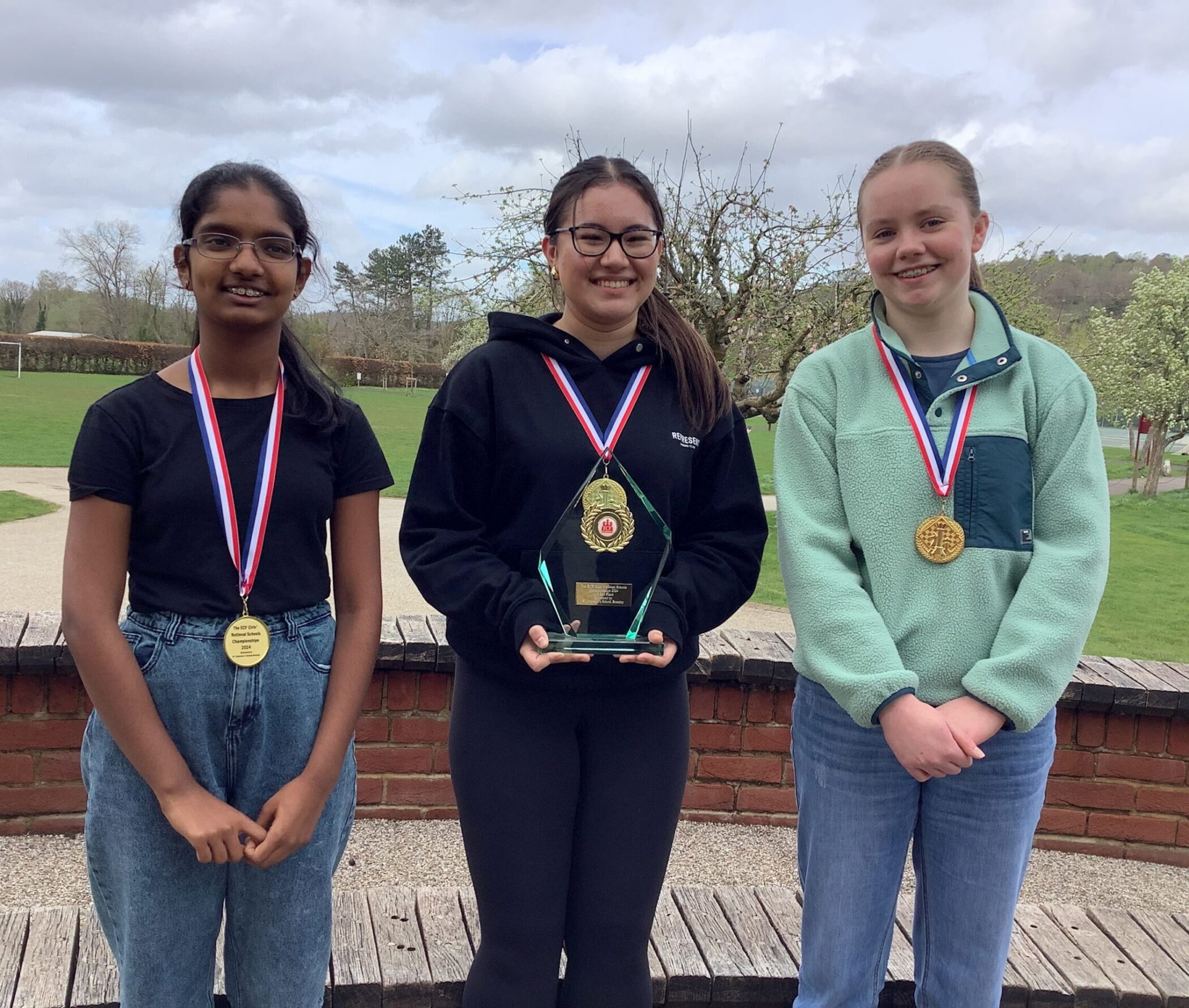 Three girls stand outdoors, smiling and wearing medals round their necks. The girl in the middle holds a trophy. Trees and grass are visible in the background under a cloudy sky.