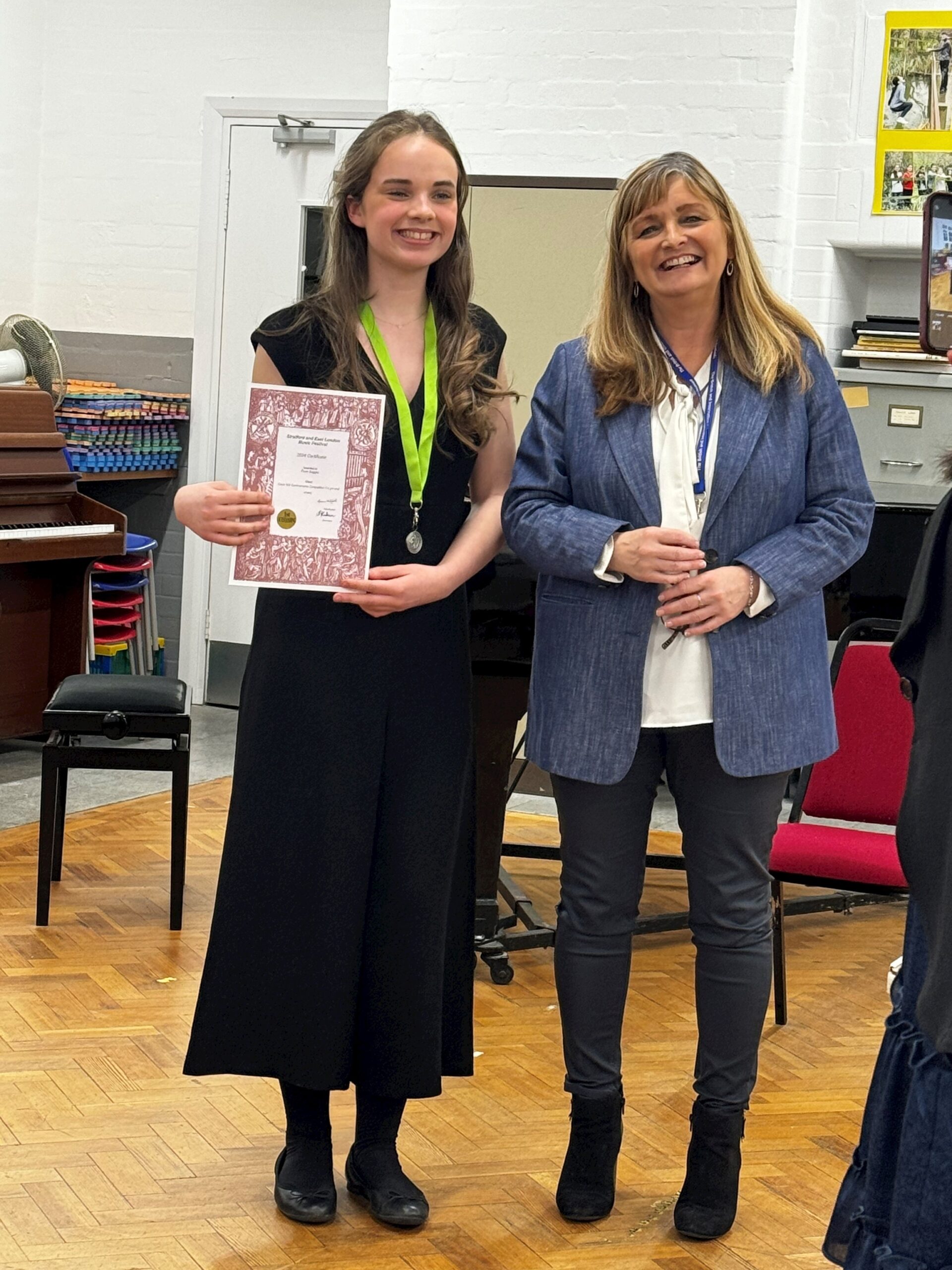 Two women stand indoors; the younger one on the left holds a certificate and wears a medal. Both are smiling. Behind them are a piano, stacked chairs, and a wall with pictures.