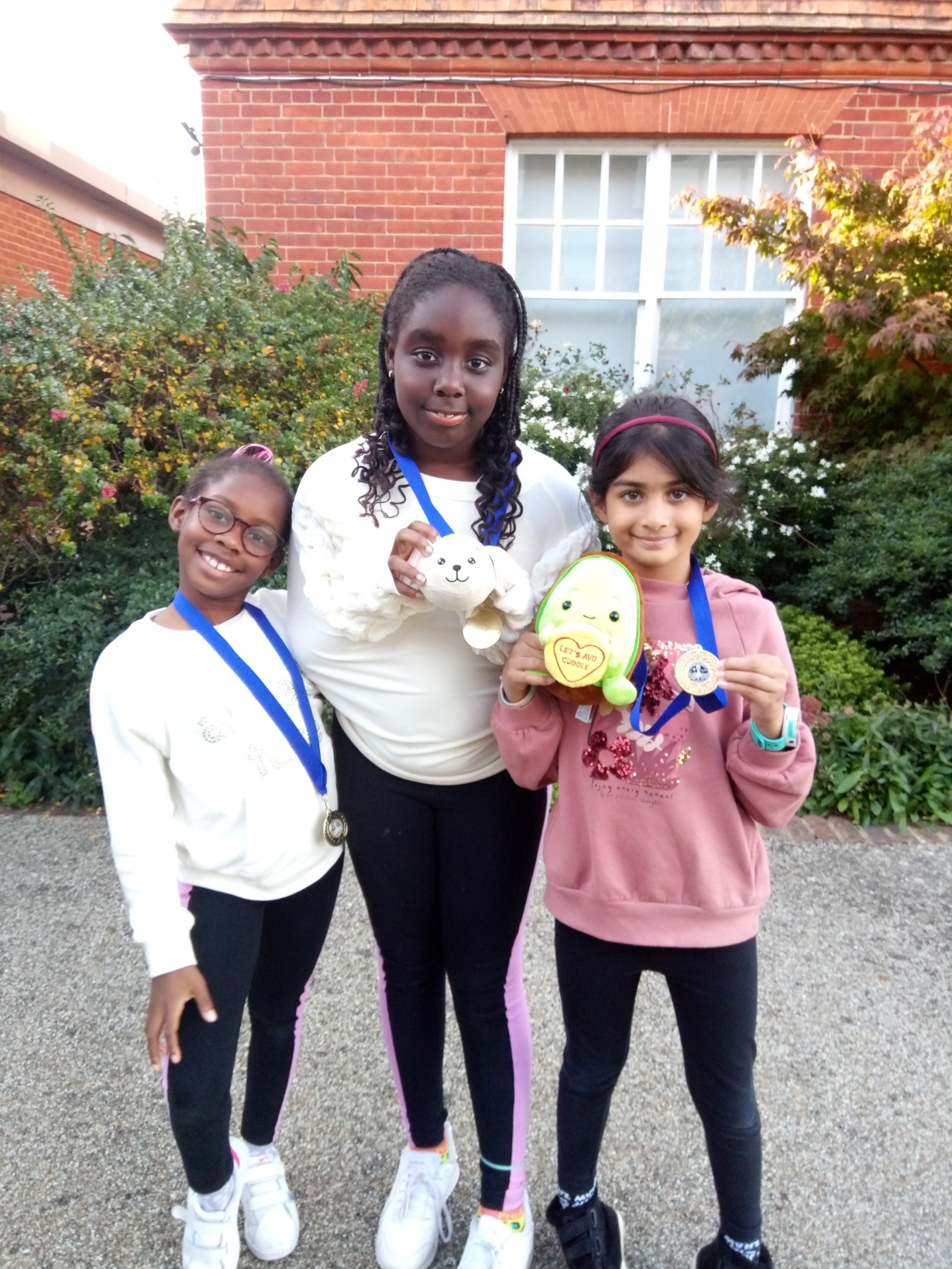 Three young girls stand outdoors, smiling and wearing medals round their necks. Two hold soft toys. They are in front of a red brick building and green foliage, posing for a cheerful photo on a sunny day.