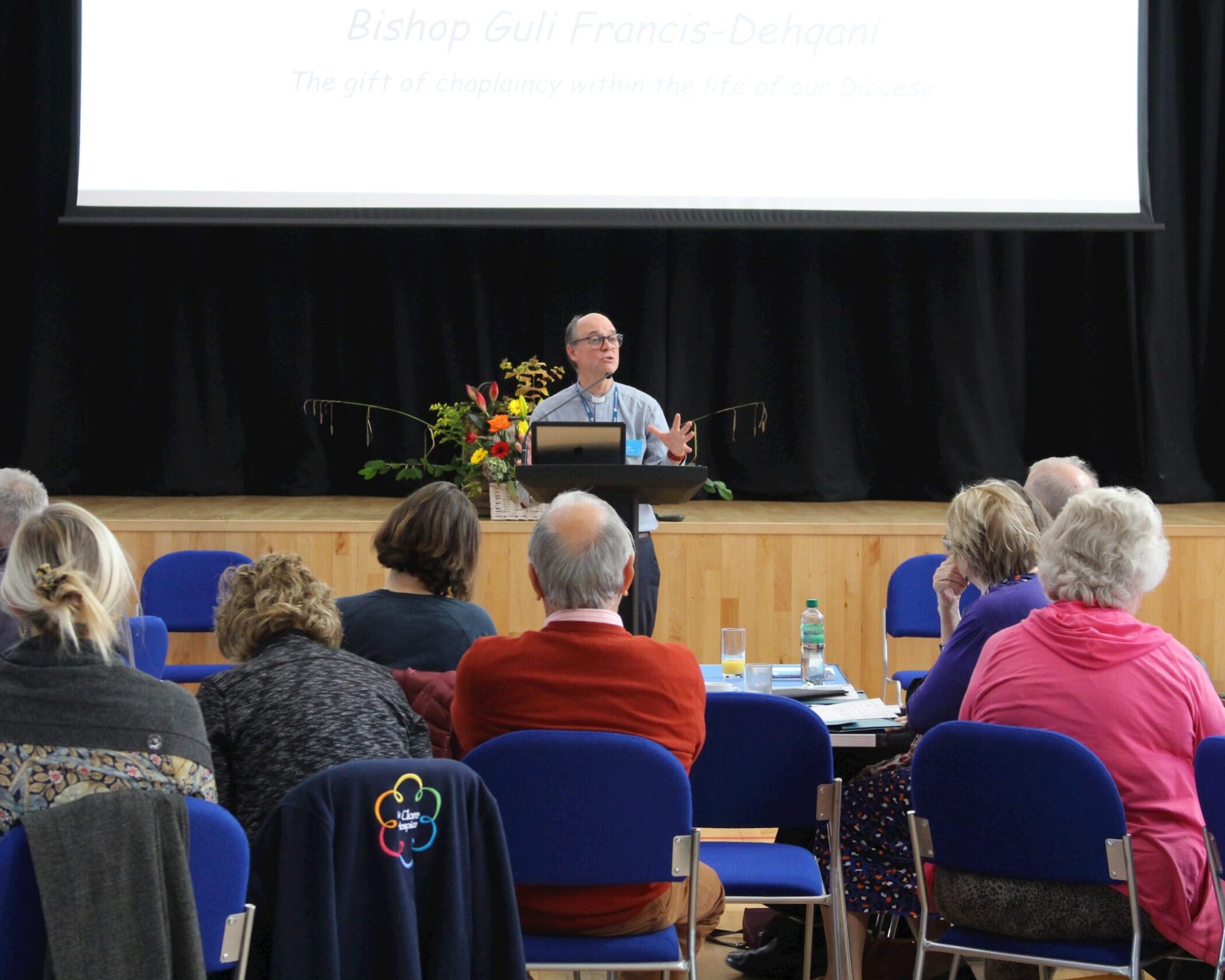 A man stands at a lectern speaking to an audience seated in blue chairs in a hall. Behind him is a large screen with text, and a flower arrangement is on the stage. The audience listens attentively.