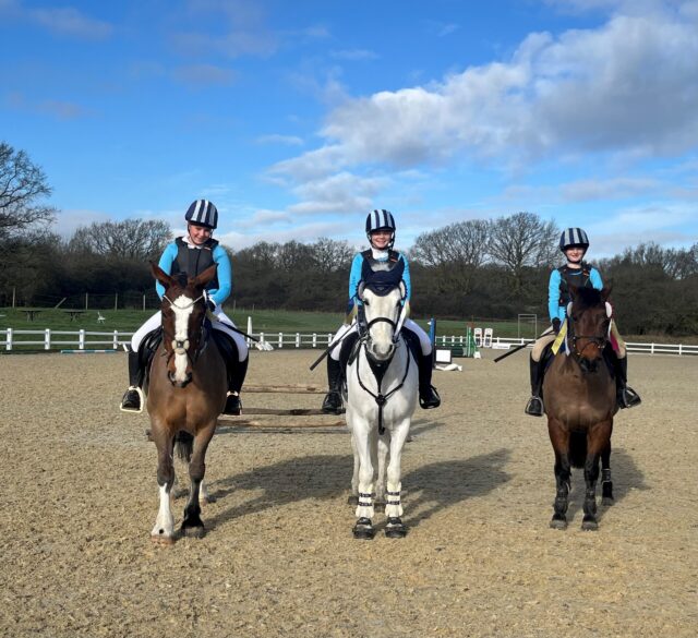 Three people wearing blue tops, black waistcoats, and striped helmets are sitting on horses in an outdoor riding arena. The sky is partly cloudy and trees line the background.