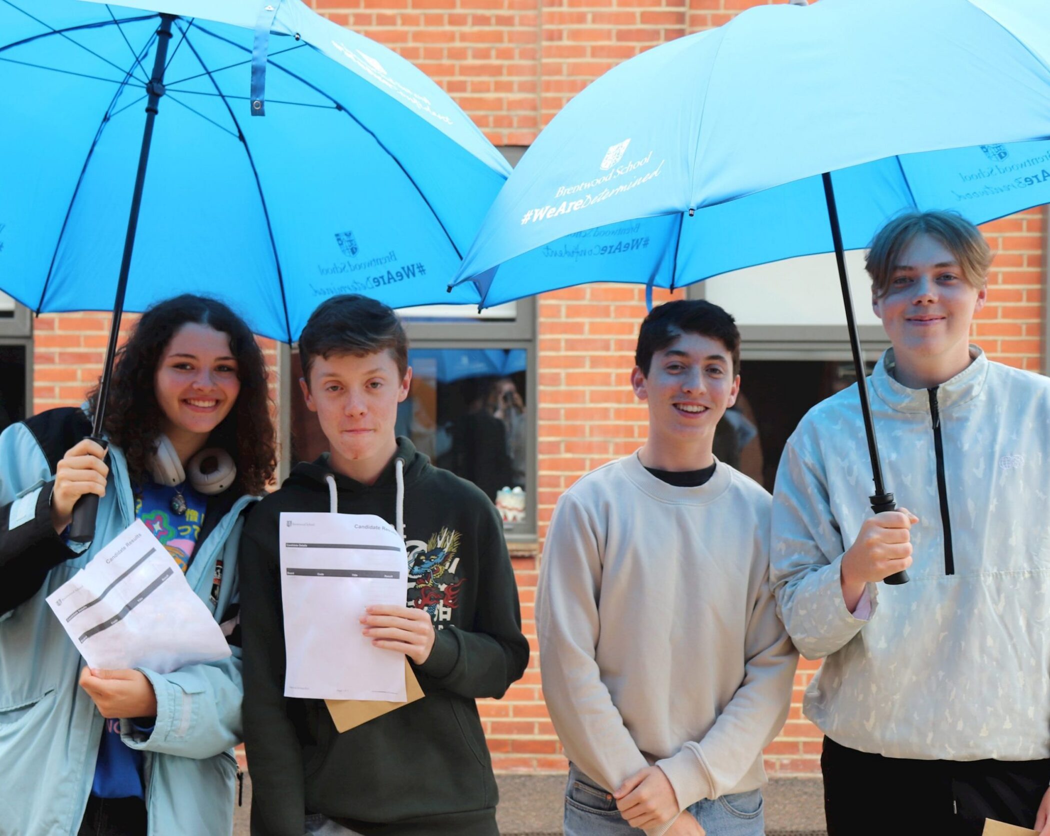 Four smiling teenagers stand outside a brick building, two holding large blue umbrellas. Three of them hold papers, and they appear happy and relaxed.
