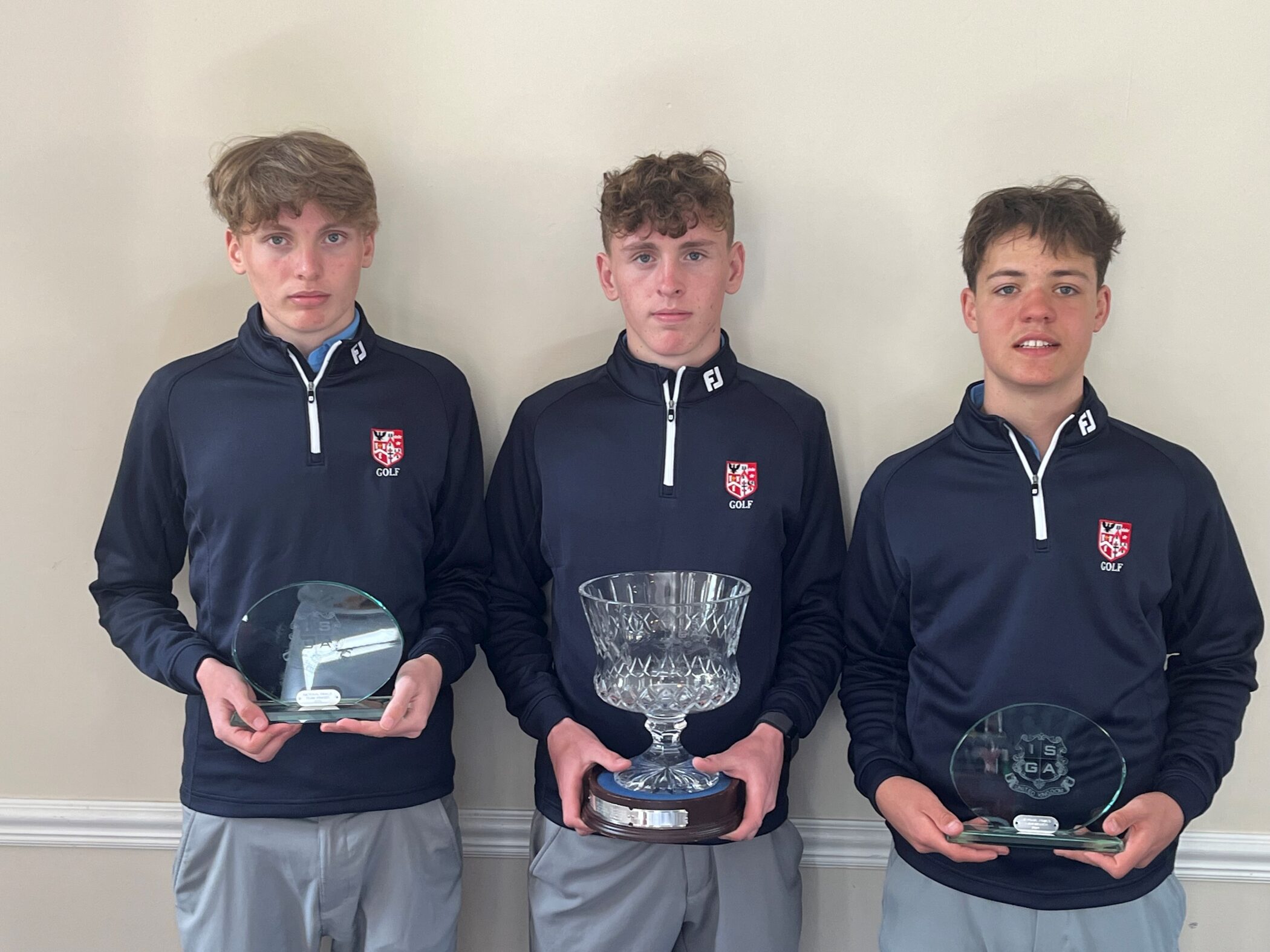 Three teenage boys in matching navy golf jackets stand side by side, each holding a trophy. The boy in the middle holds a large crystal cup, while the others hold round glass plaques. They stand against a plain beige wall.