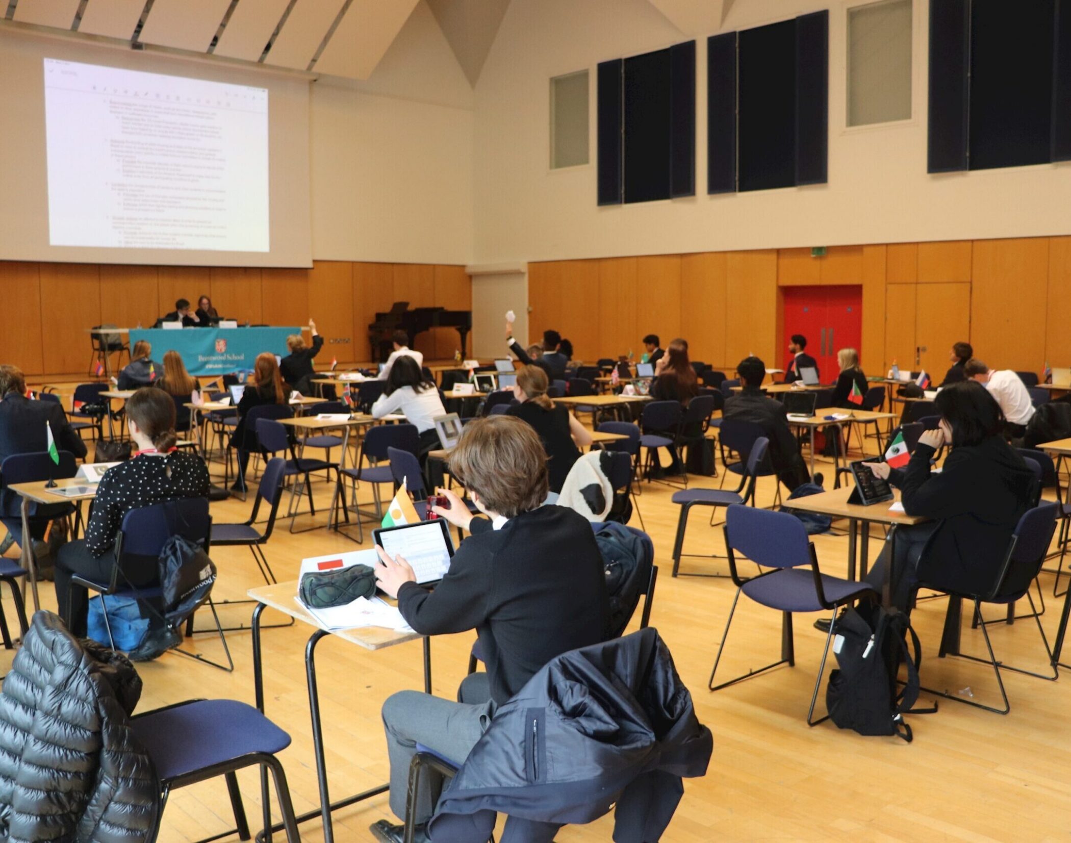 Students sit at spaced desks in a large hall, participating in a formal event or conference. Some raise their hands, while two people speak at a panel in front of a large projected document.