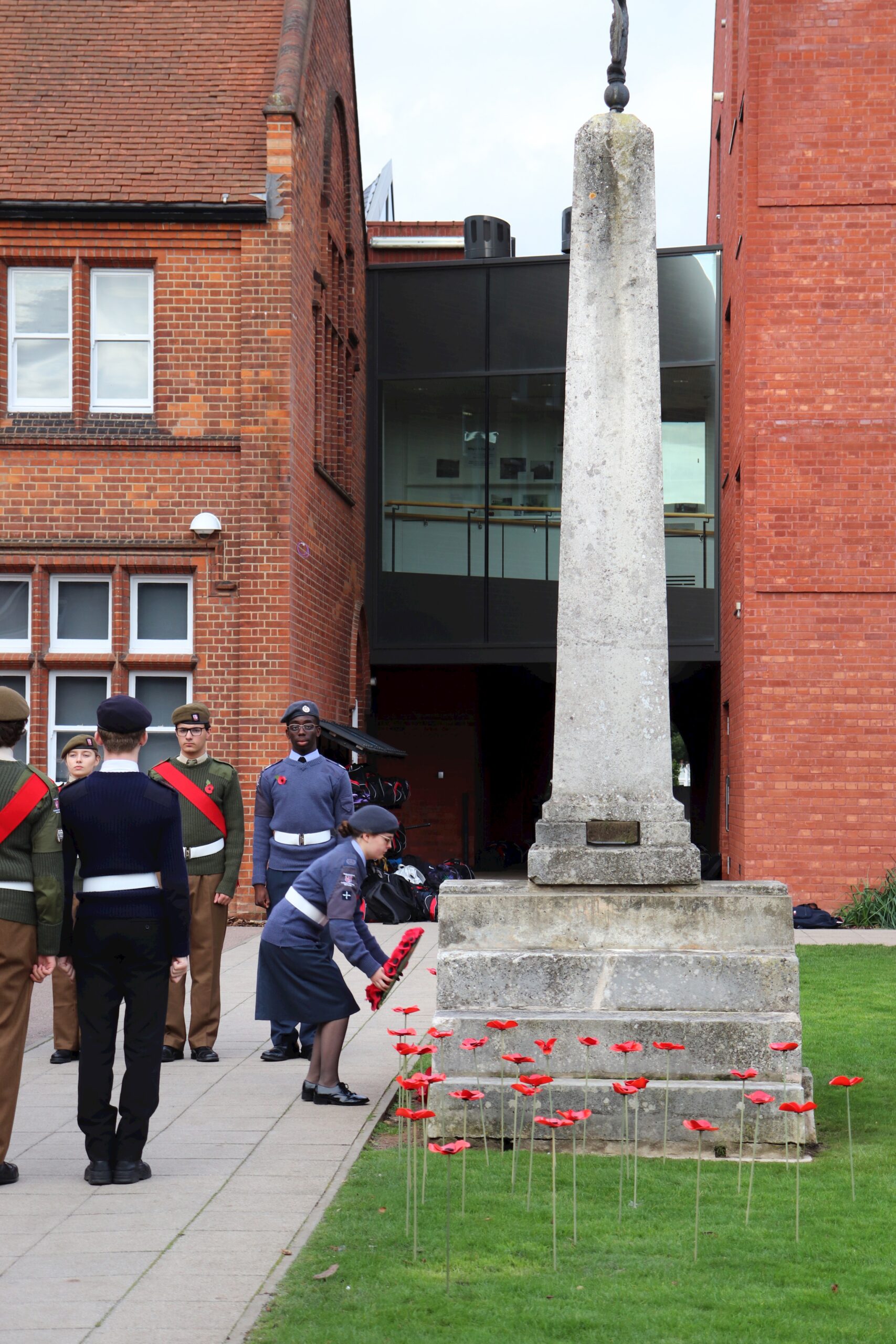 A uniformed woman places a wreath of red poppies at the base of a stone memorial outside a red brick building, as several uniformed individuals stand nearby. Red poppy decorations are arranged on the grass.